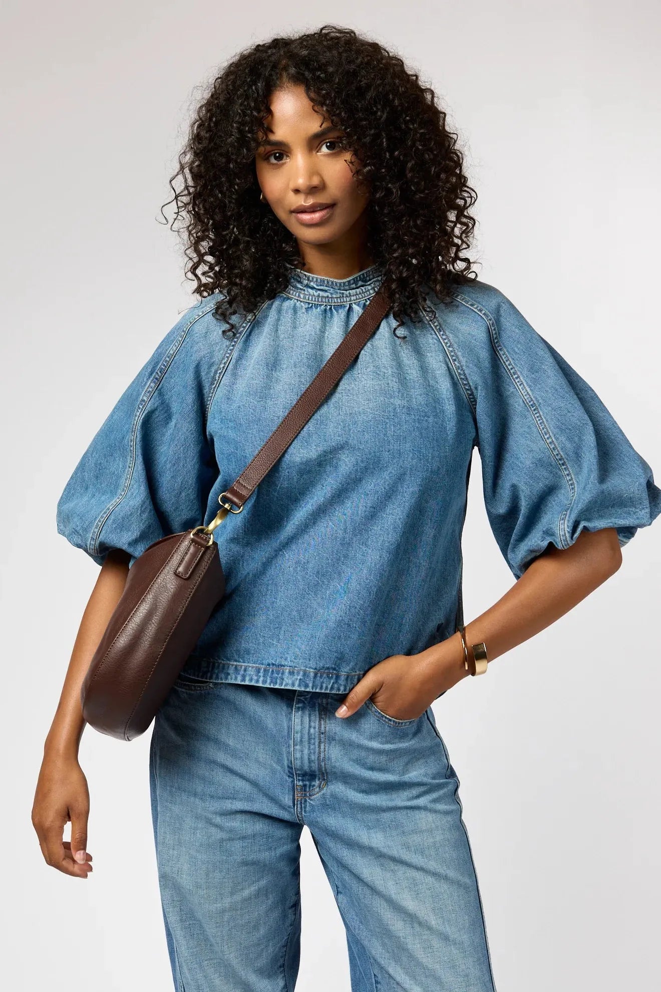 Woman wearing a denim outfit with a brown shoulder bag on a plain background