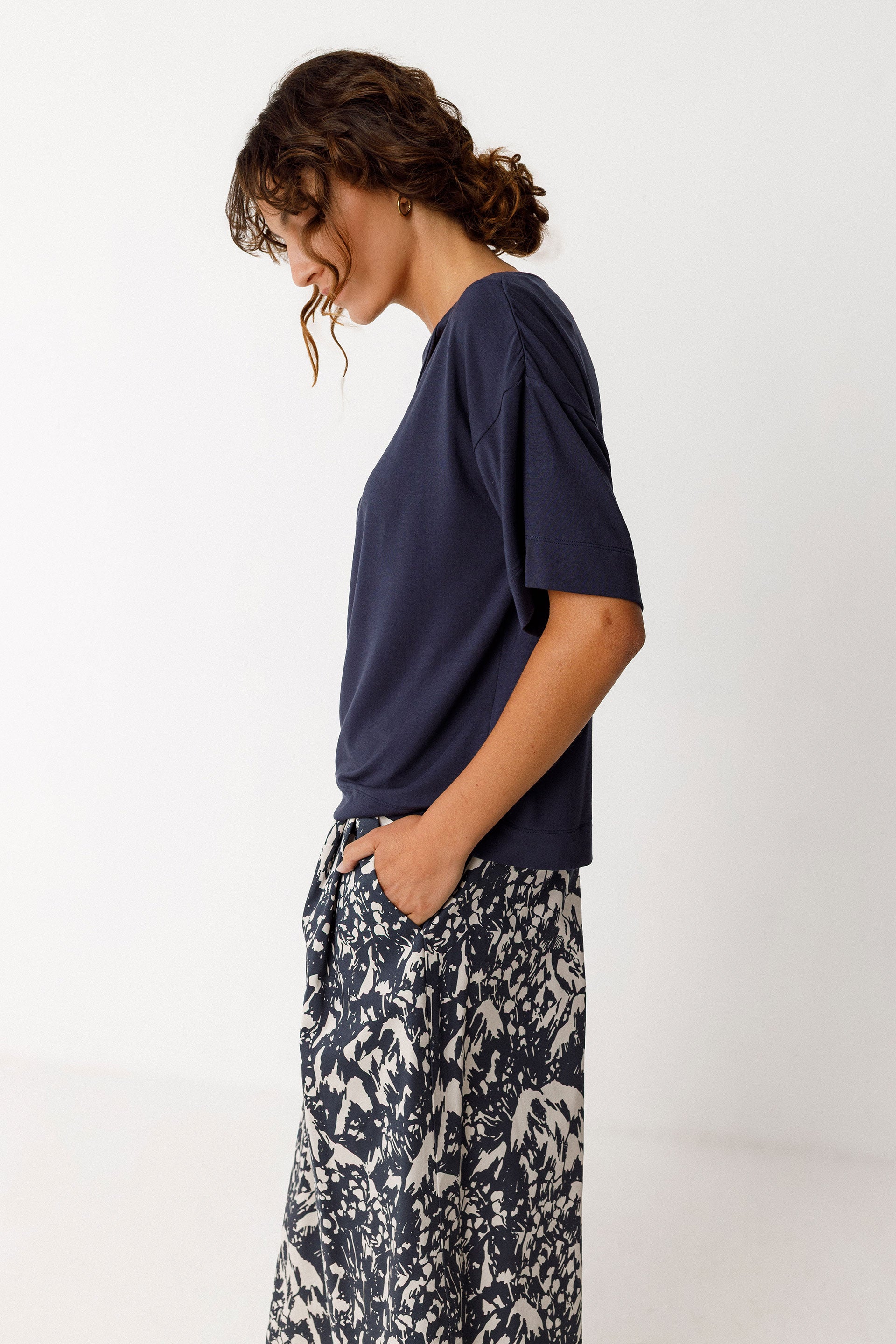 Woman wearing a loose-fitting top with a boat neck and short sleeves, featuring a wrapped effect at the front and patterned pants on a white background