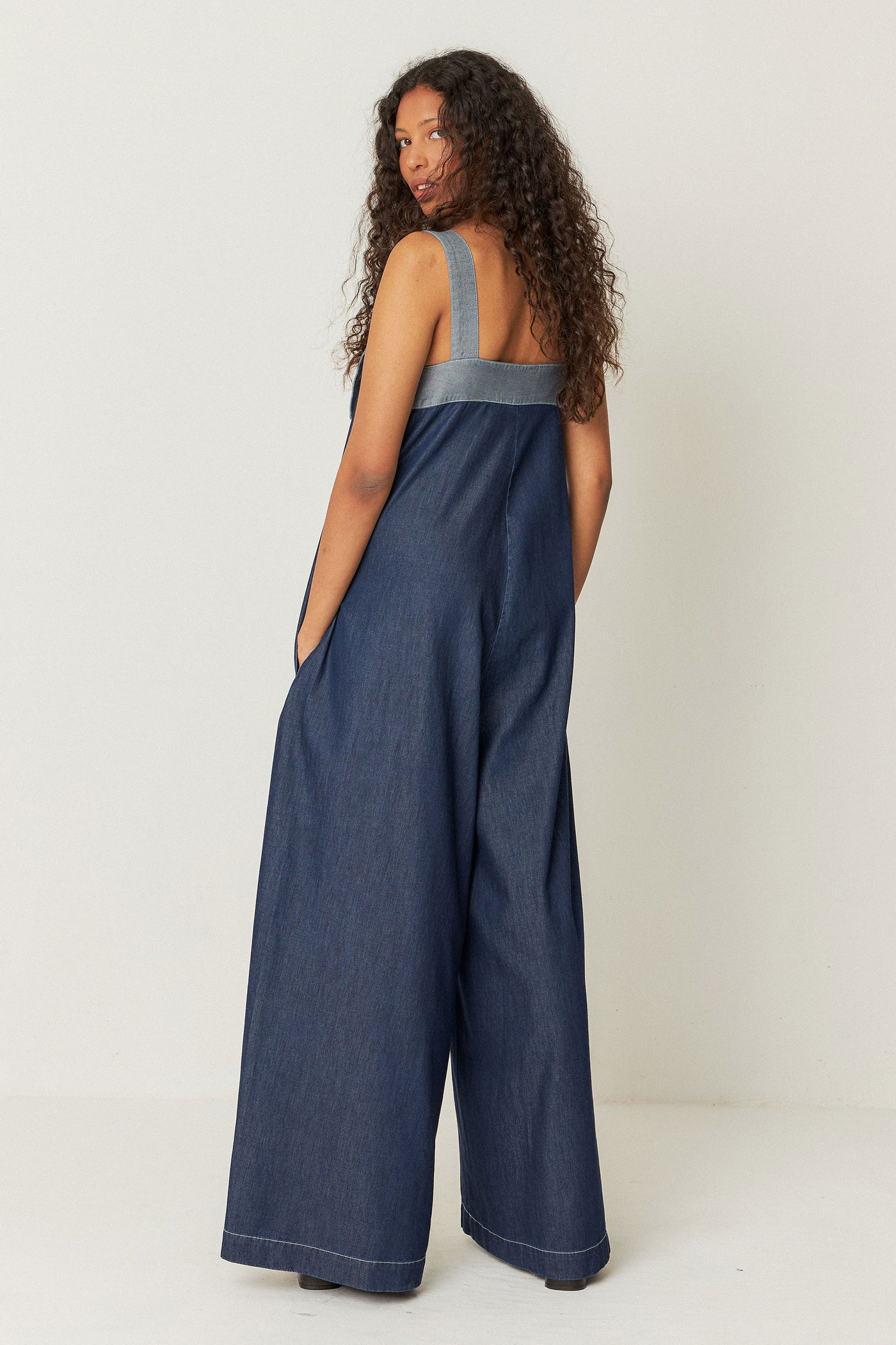 back view of model wearing long, relaxed-fit denim jumpsuit featuring front pockets, decorative stitching, and a zip closure