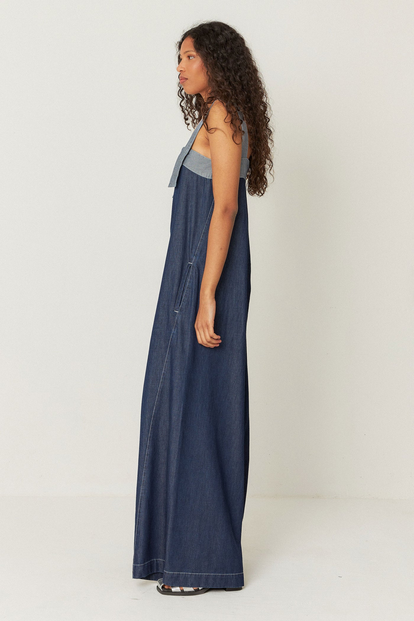 side view of model wearing long, relaxed-fit denim jumpsuit featuring front pockets, decorative stitching, and a zip closure