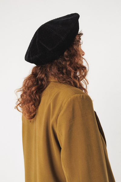 Person wearing a black organic cotton beret and mustard-colored coat on a white background