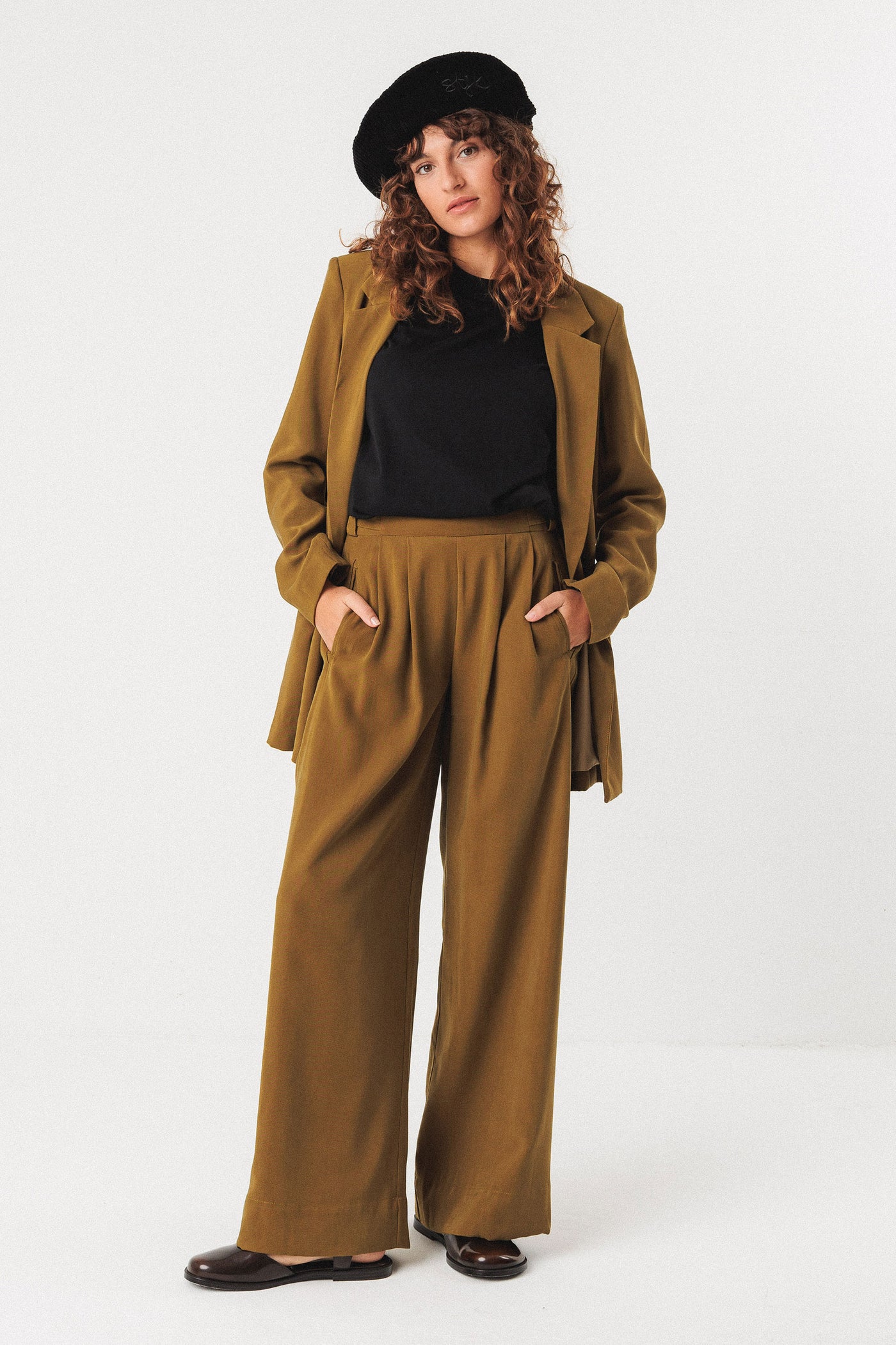 Woman wearing a brown coat and pants with a black organic cotton beret on a white background