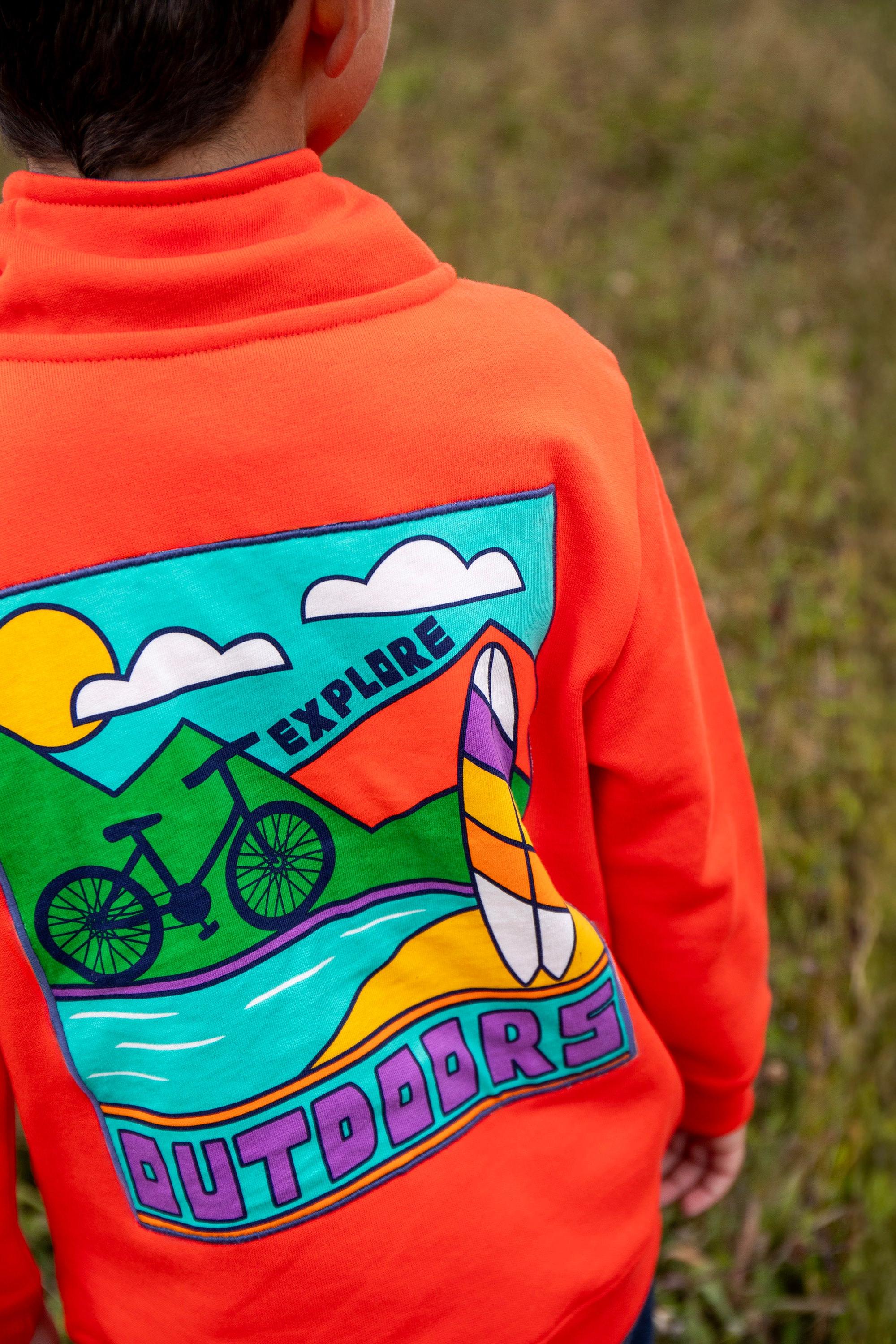 Child wearing an orange hoodie with a colorful 'Explore Outdoors' graphic on the back, standing in a grassy field.