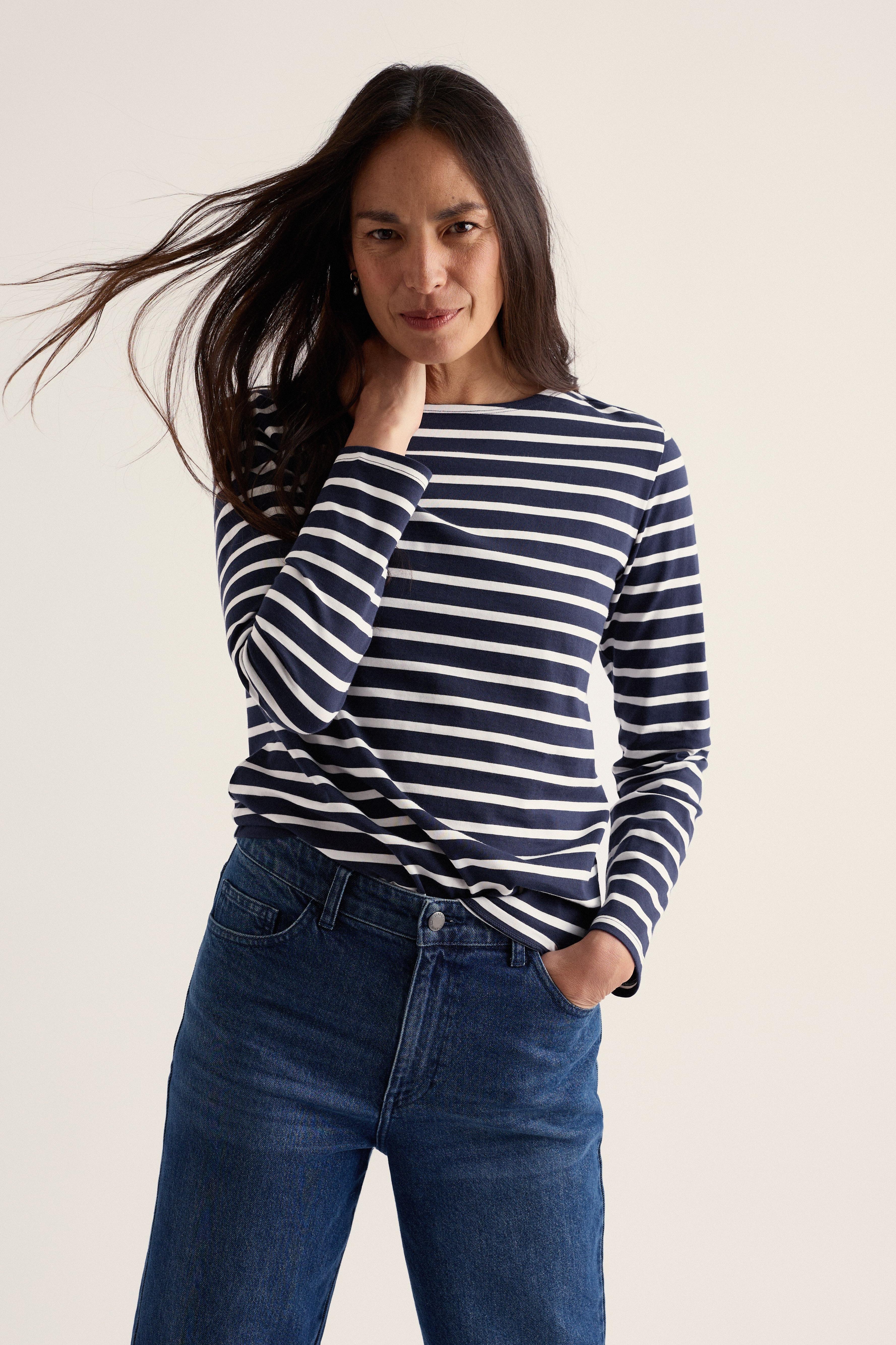 A woman modeling a navy and white horizontally striped long-sleeve top with dark blue jeans, set against a plain light beige background.