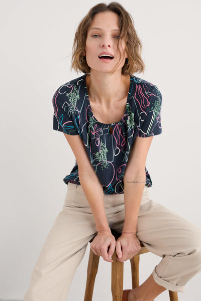 Woman sitting on a wooden stool smiling, wearing the Appletree T-shirt with cream trousers.