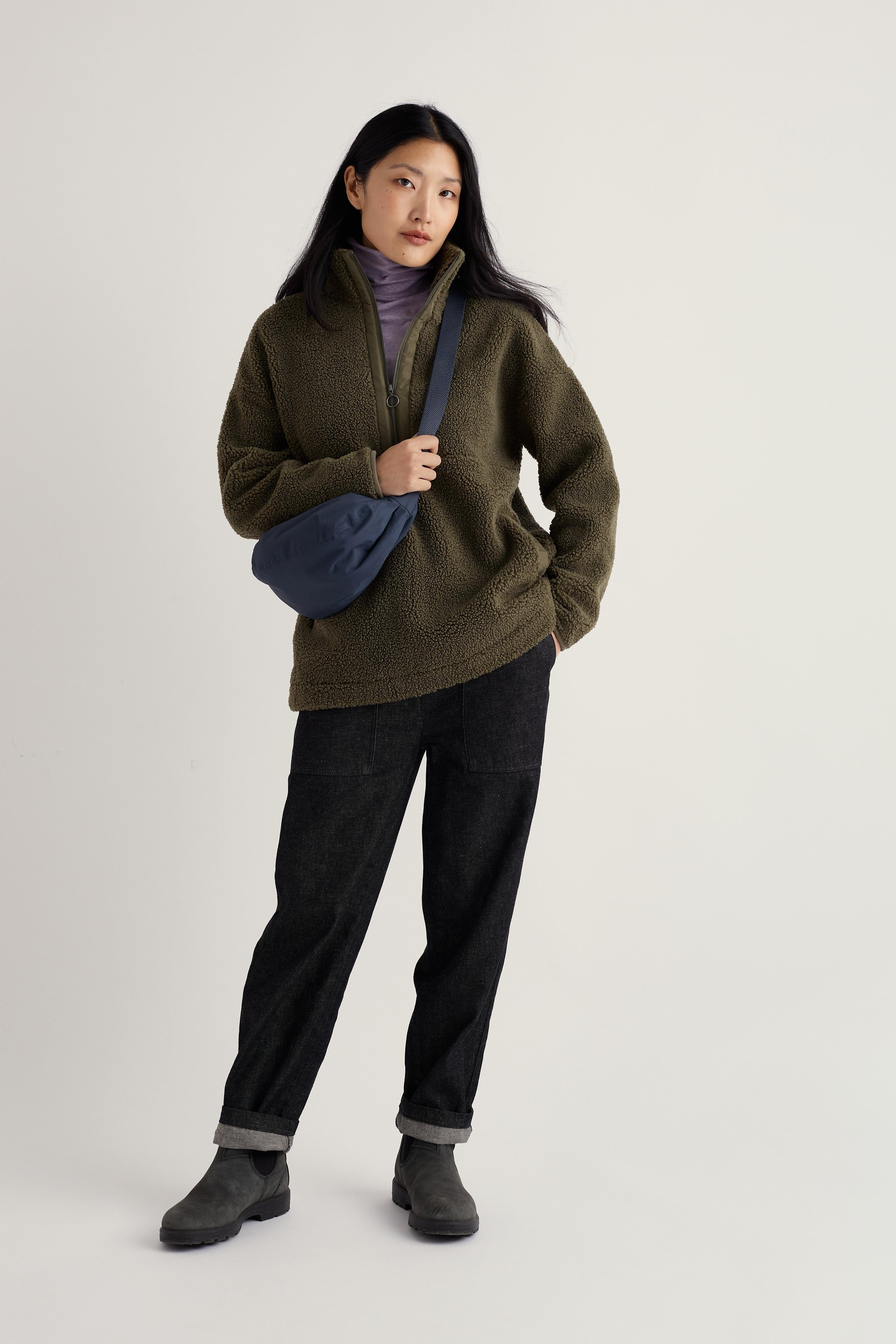 Person wearing a green sweater and dark pants holding a blue bag on a light gray background