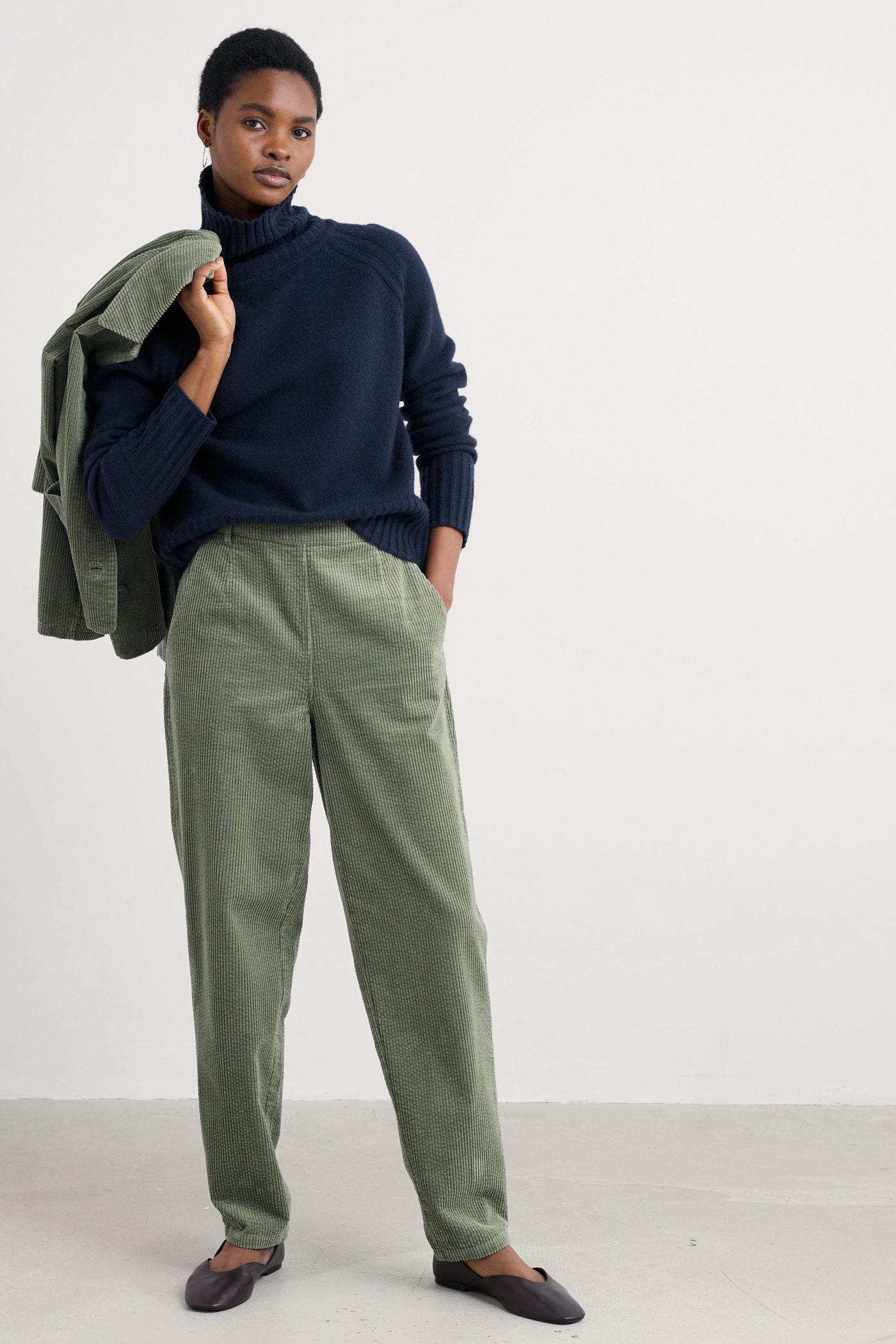 Person wearing a navy wool sweater and green pants holding a green jacket against a white background