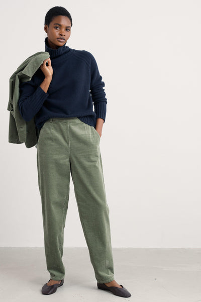 Person wearing a navy wool sweater and green pants holding a green jacket against a white background