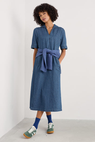 Model wearing the Seasalt Fen Violet Denim Dress in mid-wash denim, featuring a V-neck with a nautical-style collar, short sleeves, and a fitted waist leading to a full, midi-length skirt.
