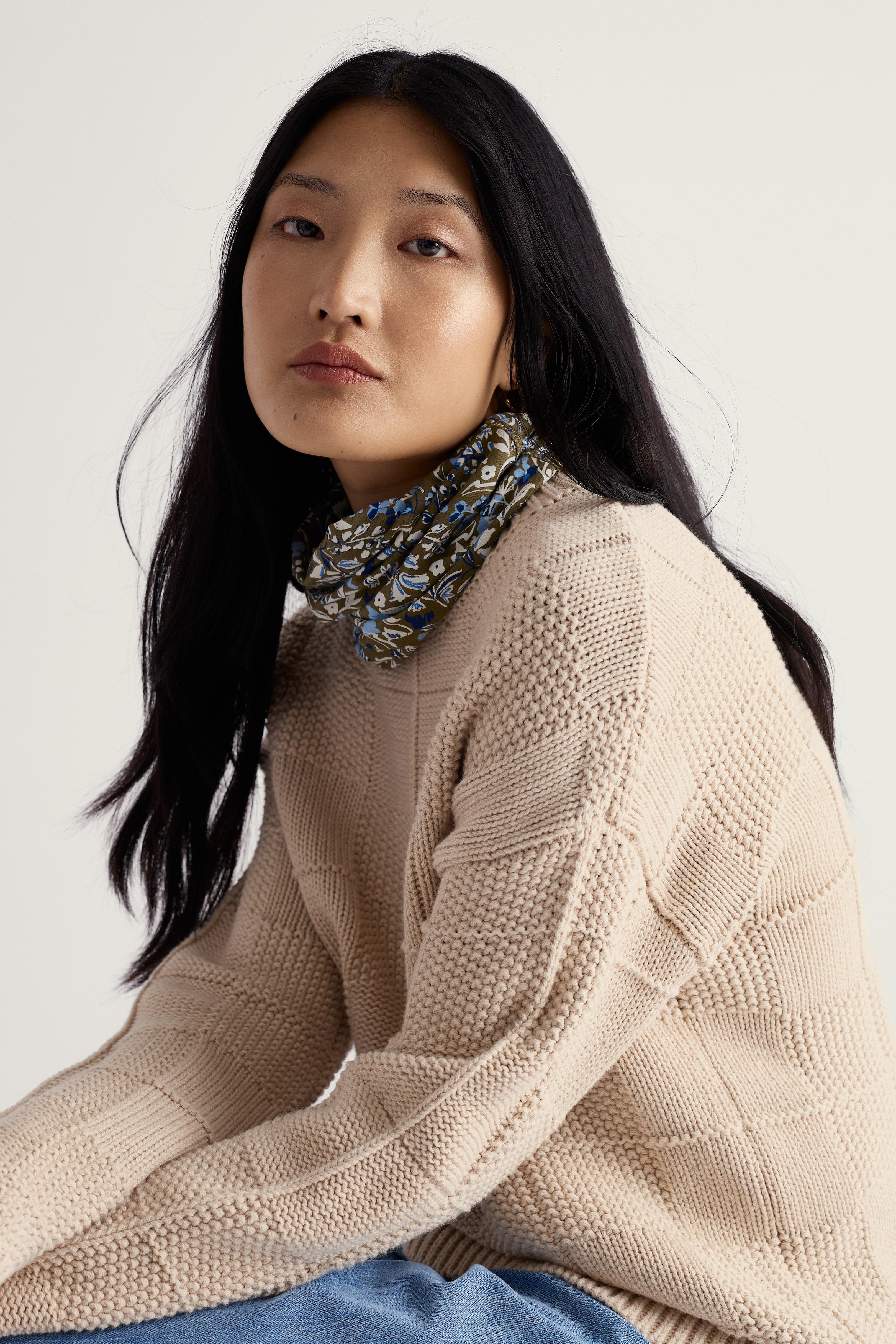 Woman wearing a beige knitted sweater with a patterned scarf on a plain background