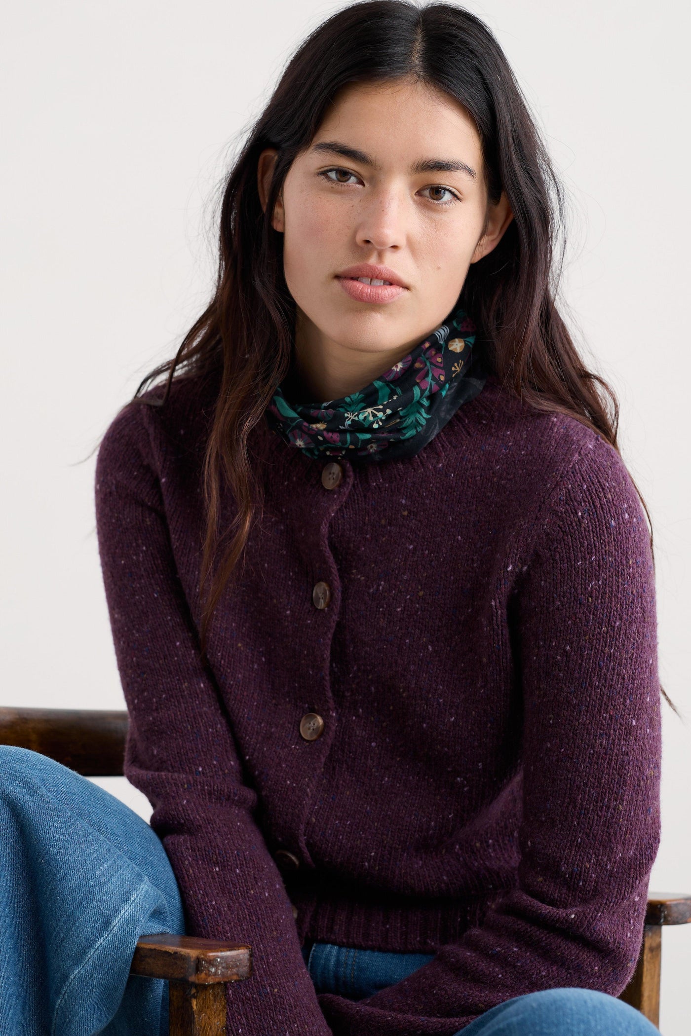 Woman wearing a purple wool sweater with a floral scarf, sitting on a wooden chair against a white background.