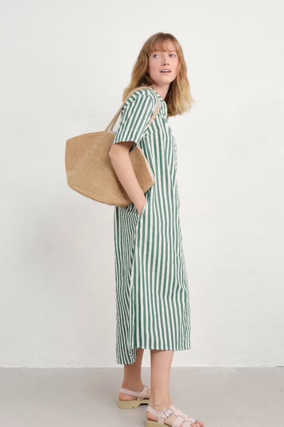 Side view of model wearing green striped Harbour Movement Dress, hands in pockets, showing relaxed fit and curved hem.