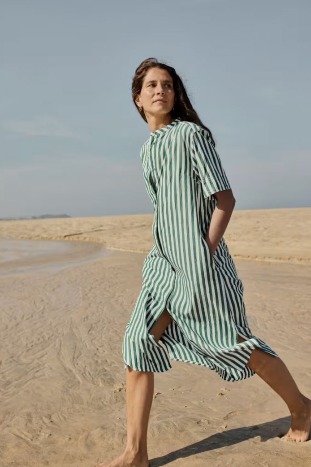 Woman walking barefoot on a sandy beach wearing a Seasalt midi-length shirt dress with green and white vertical stripes.