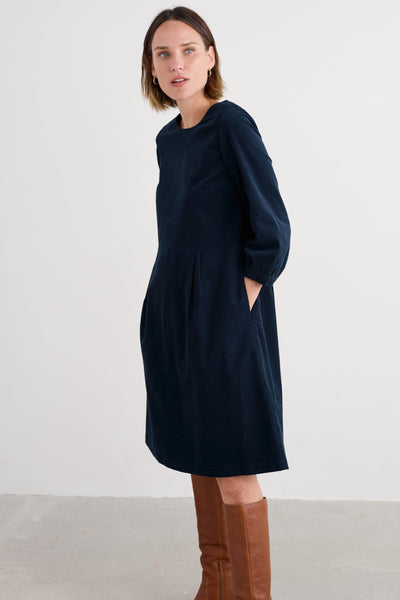 Woman wearing a navy blue organic cotton dress with brown knee-high boots on a white background