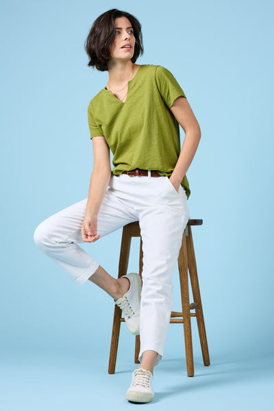 Woman sitting sideways on a stool wearing the Dark Citron Seasalt T-Shirt and white trousers, casual and relaxed pose.