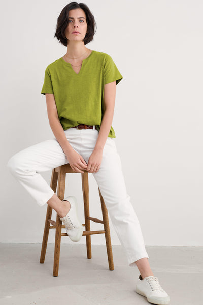 Full front view of woman wearing Seasalt Kellow T-Shirt in Dark Citron, styled with white trousers and white trainers.
