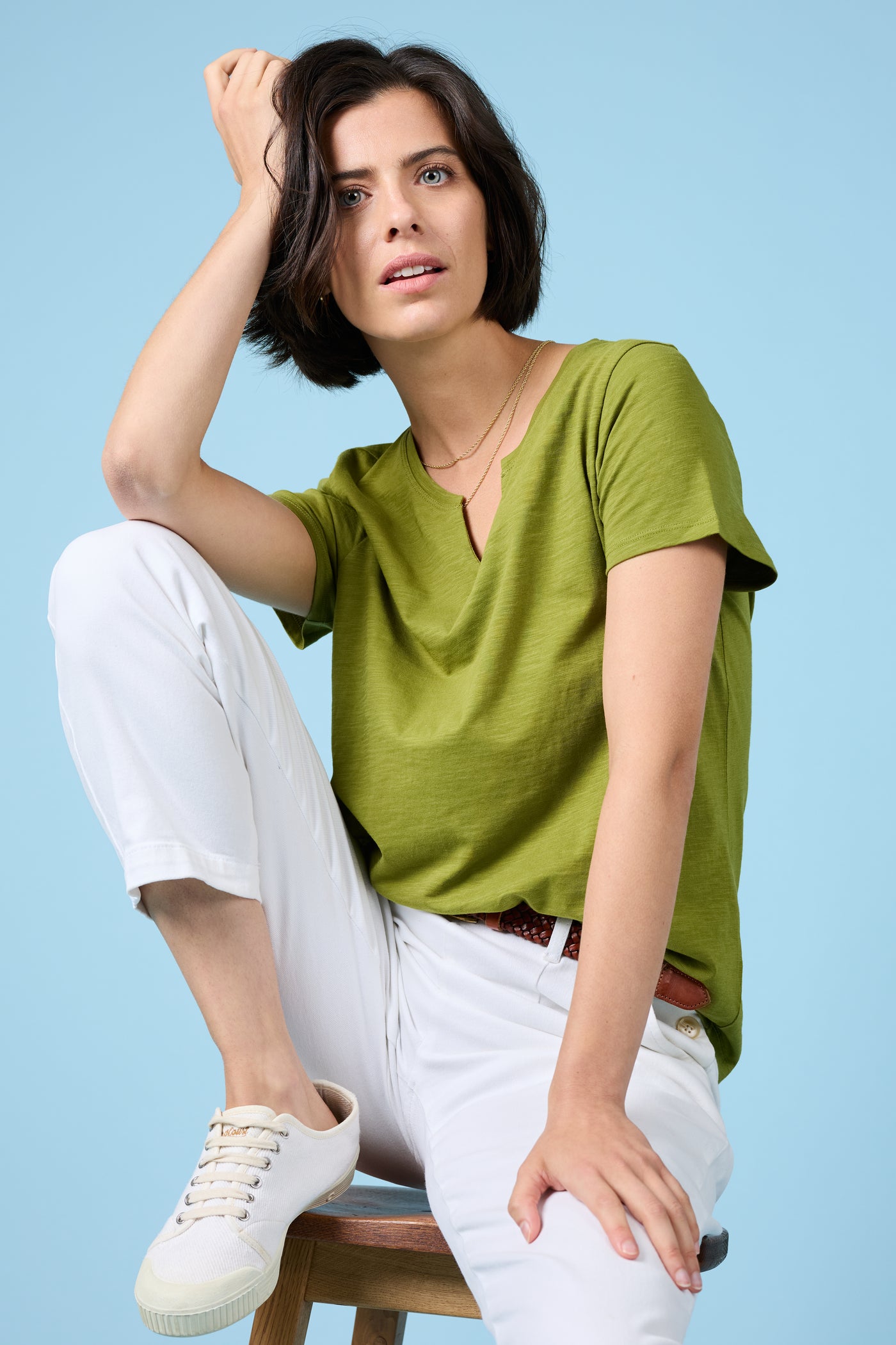 Woman wearing the Seasalt Kellow T-Shirt in Dark Citron, seated on a stool with white trousers and trainers, against a light blue background.