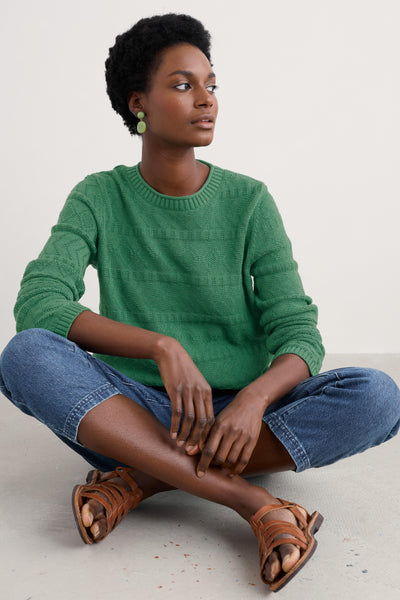 Model sitting with folded legs wearing organic cotton knit jumper paired with denim jeans.