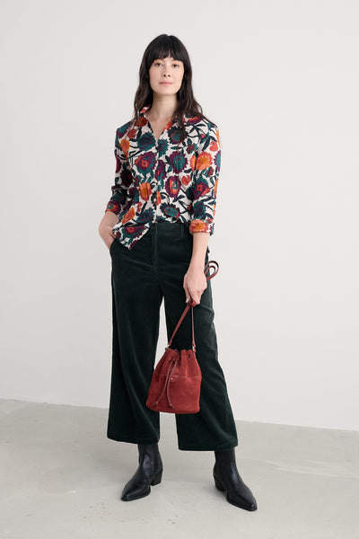 Woman wearing a floral organic cotton blouse with green cord pants and holding a red bag against a white background