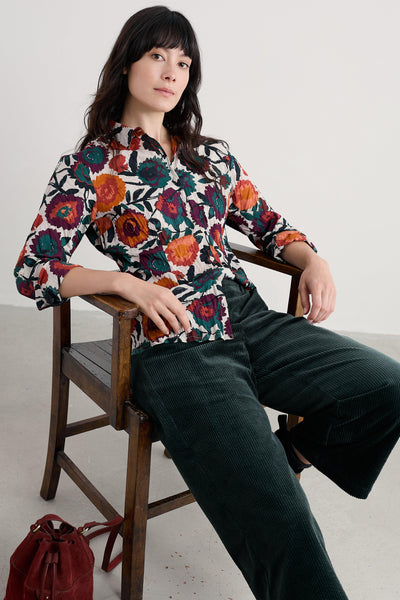 Woman sitting on a wooden chair wearing a colorful floral blouse and dark green pants.