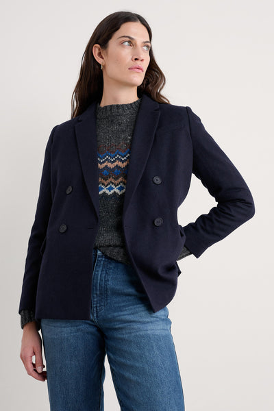 Woman wearing a navy blazer, gray sweater with pattern, and blue jeans on a light background