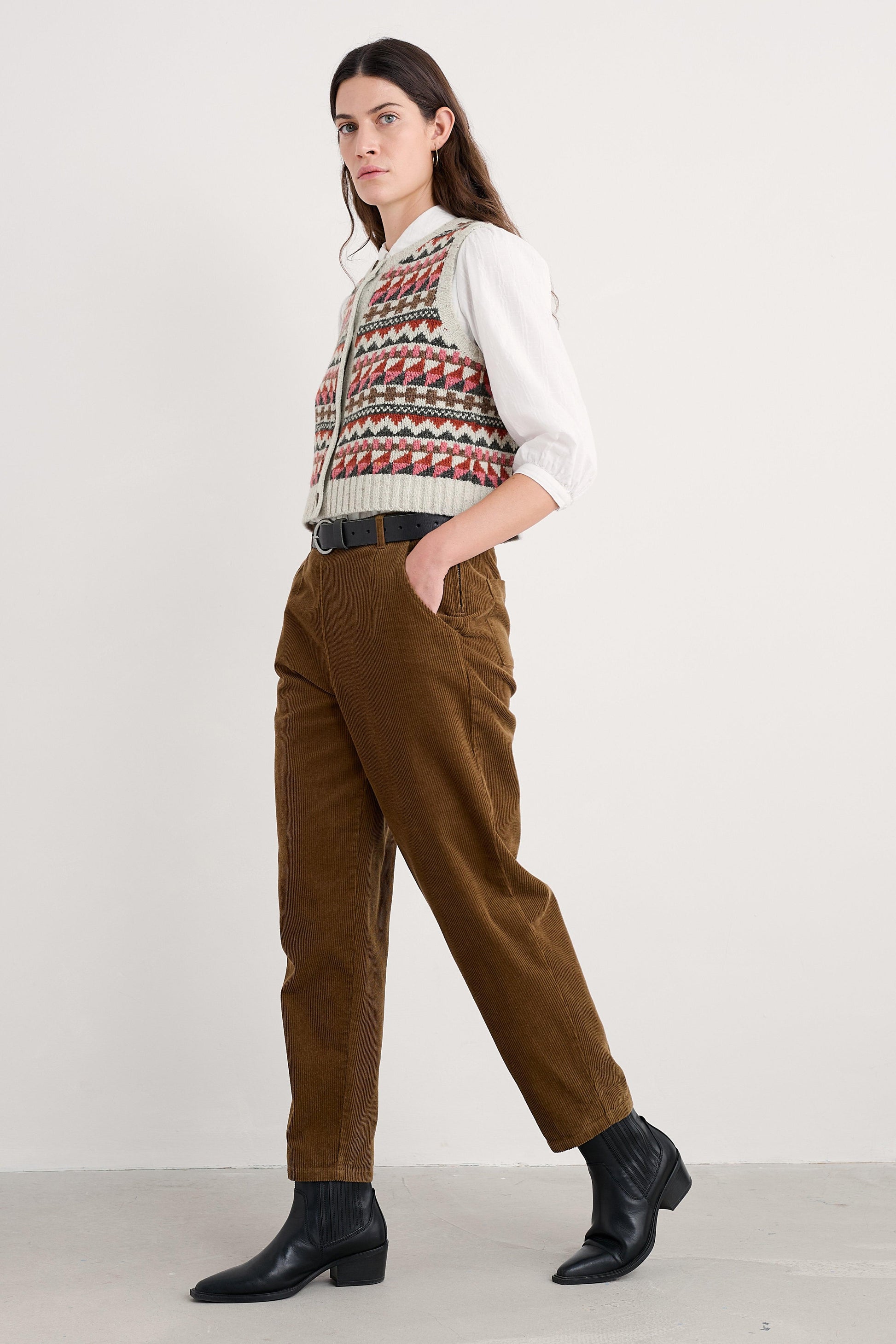 Woman wearing a patterned vest, white shirt, brown pants, and black shoes on a white background