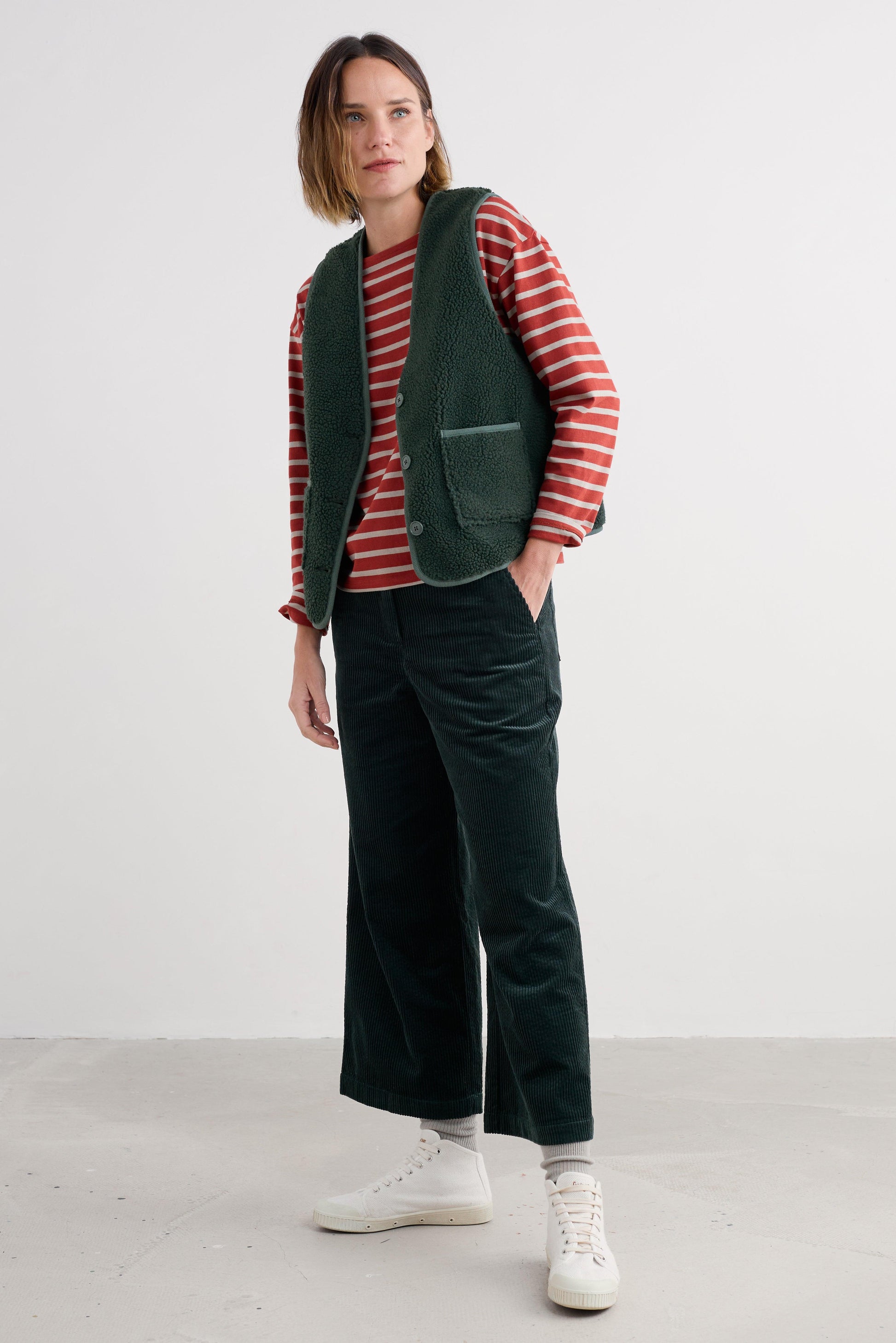 Woman wearing a red striped shirt, green vest, and dark green pants on a white background