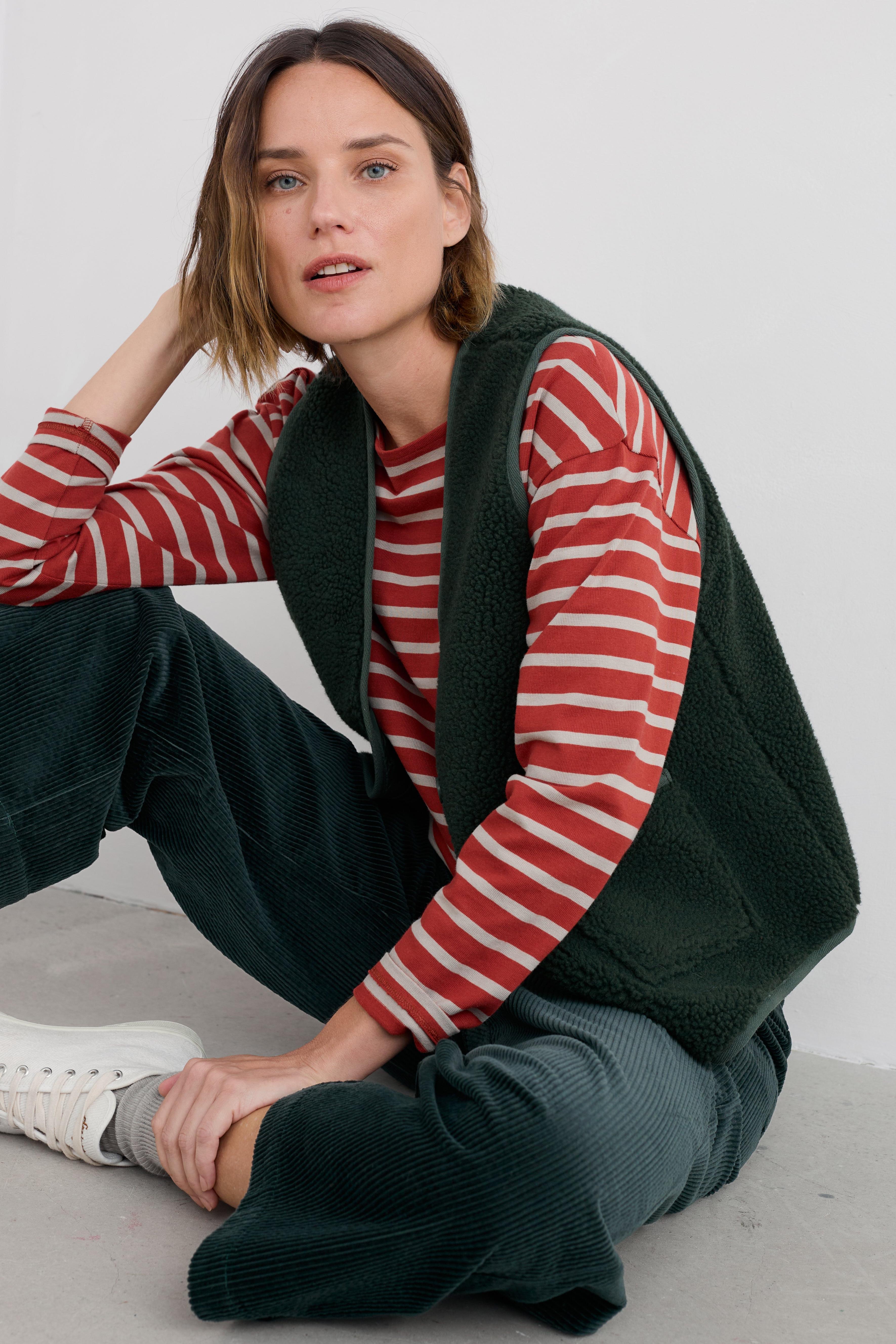 Woman wearing a red and white striped sweater with a recycled green vest sitting on a light-colored floor.