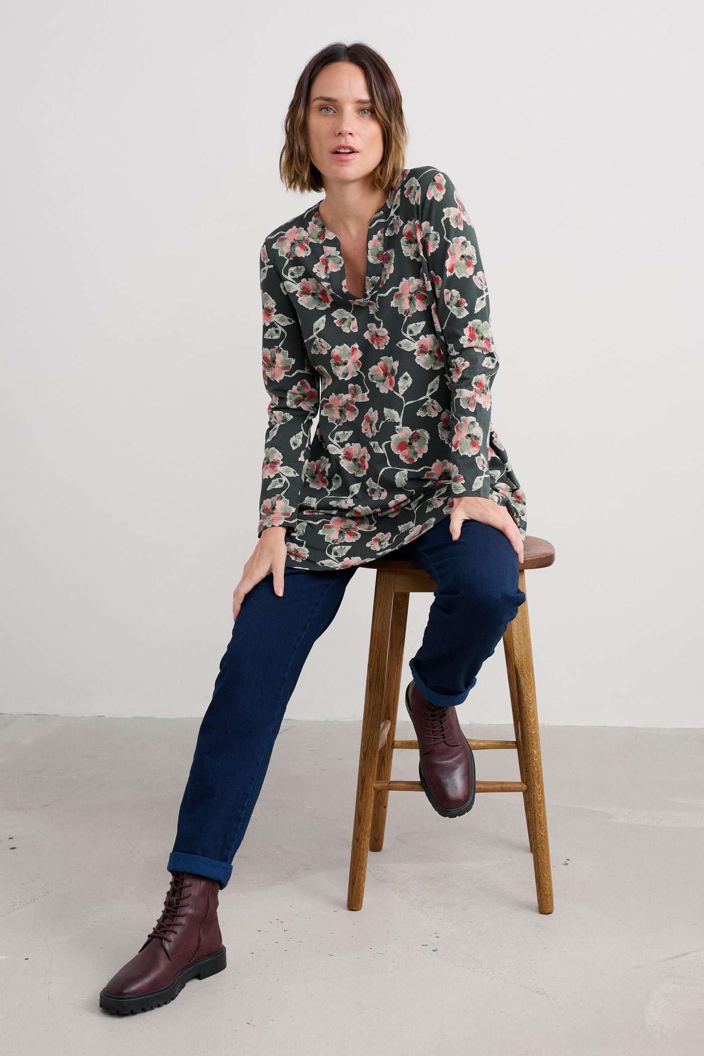 Woman sitting on a stool wearing a floral organic cotton tunic and dark blue jeans with brown boots.
