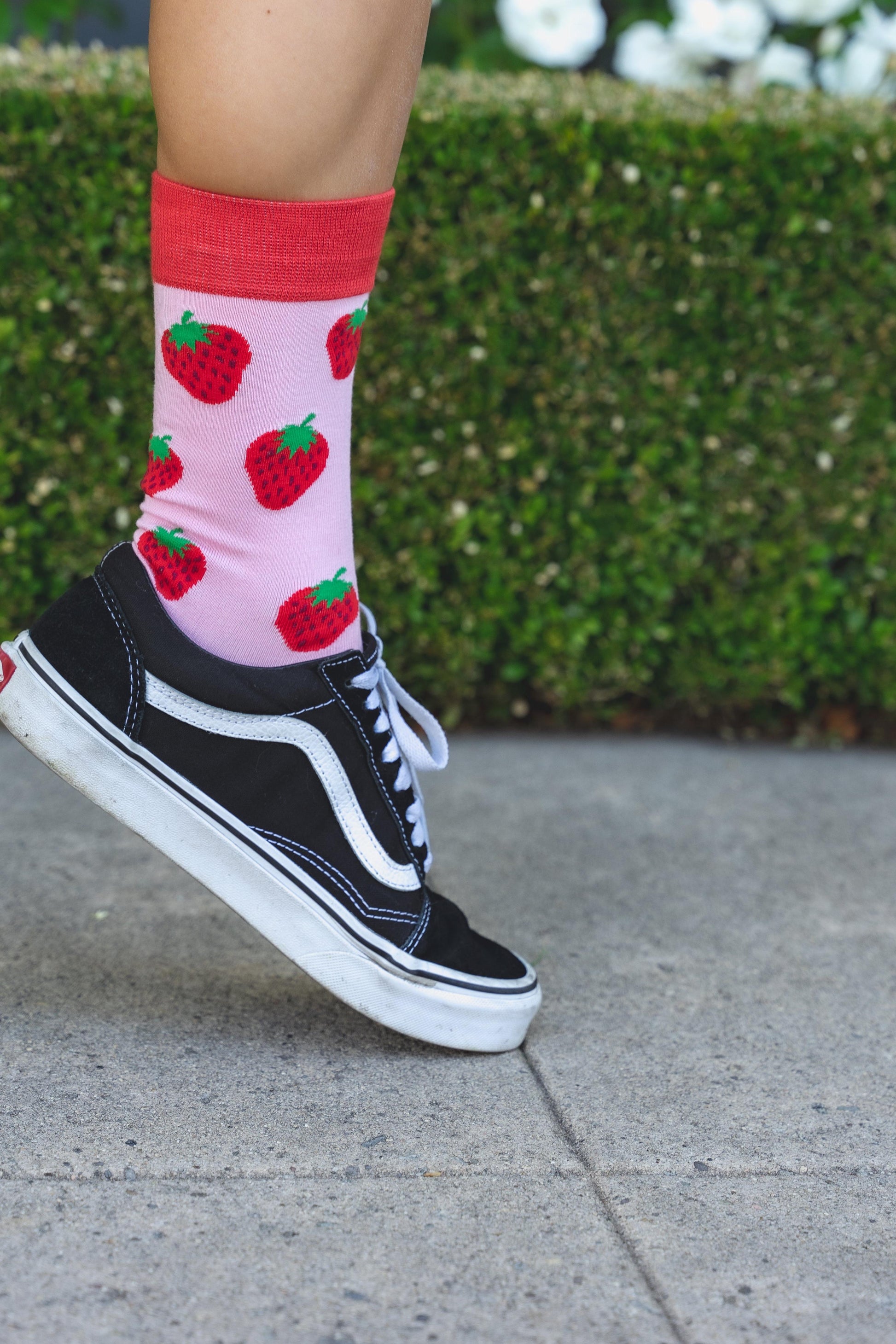 Person wearing black Vans sneakers with pink socks featuring red strawberry pattern on a pavement background.