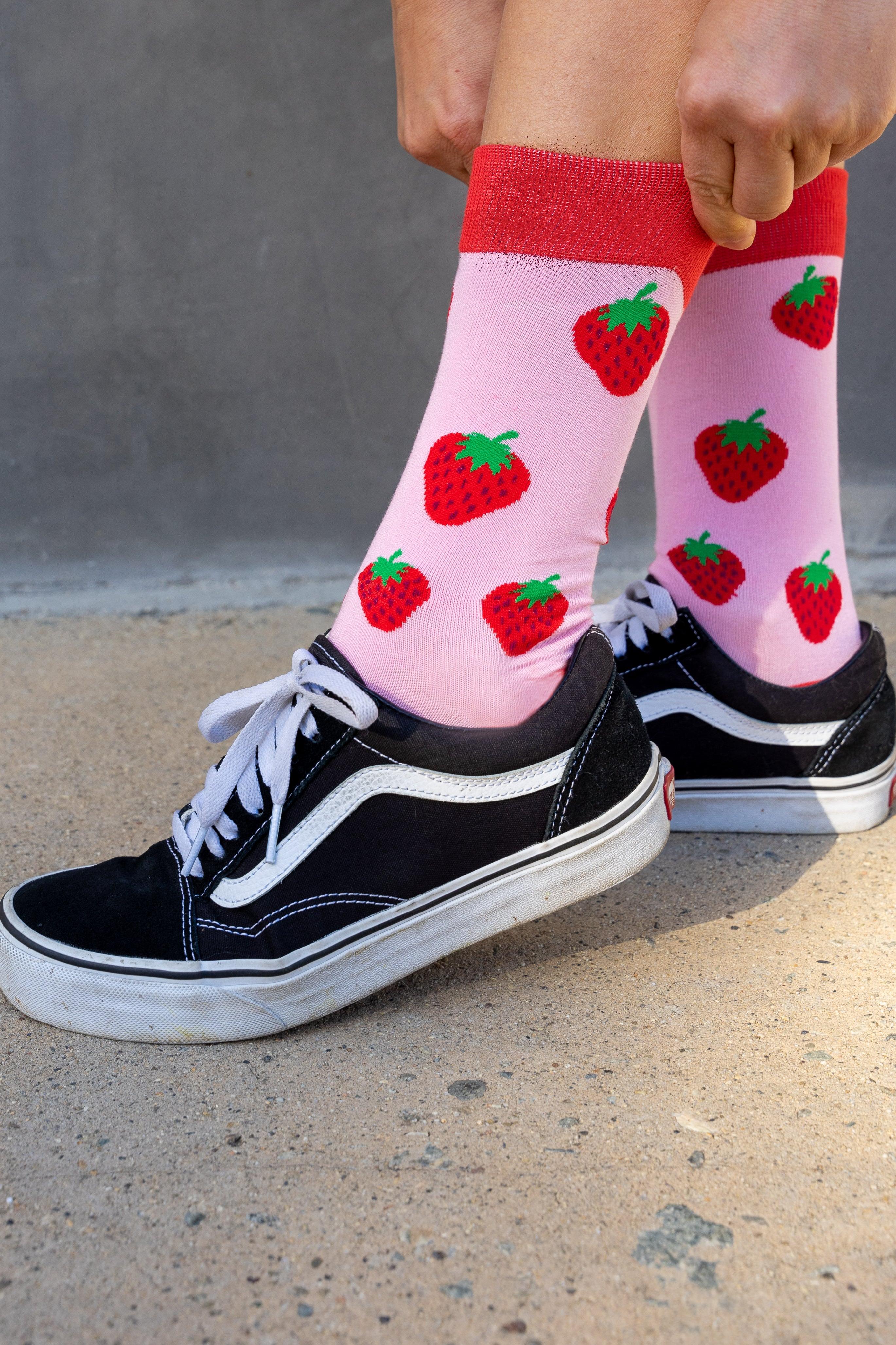 Person wearing pink bamboo socks with strawberry pattern and black sneakers on a concrete floor.