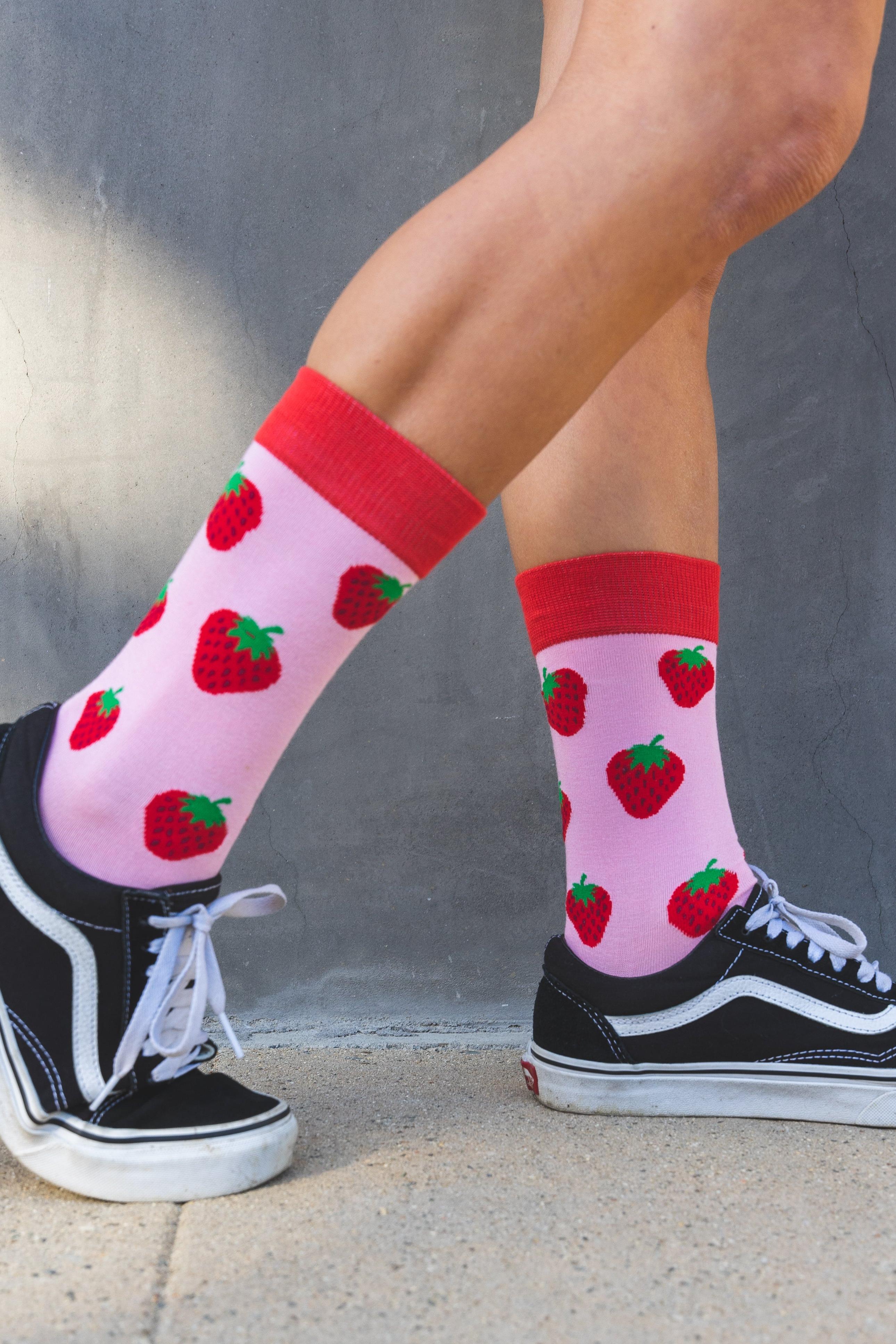 Person wearing pink socks with red strawberry pattern and black Vans sneakers against a gray wall.