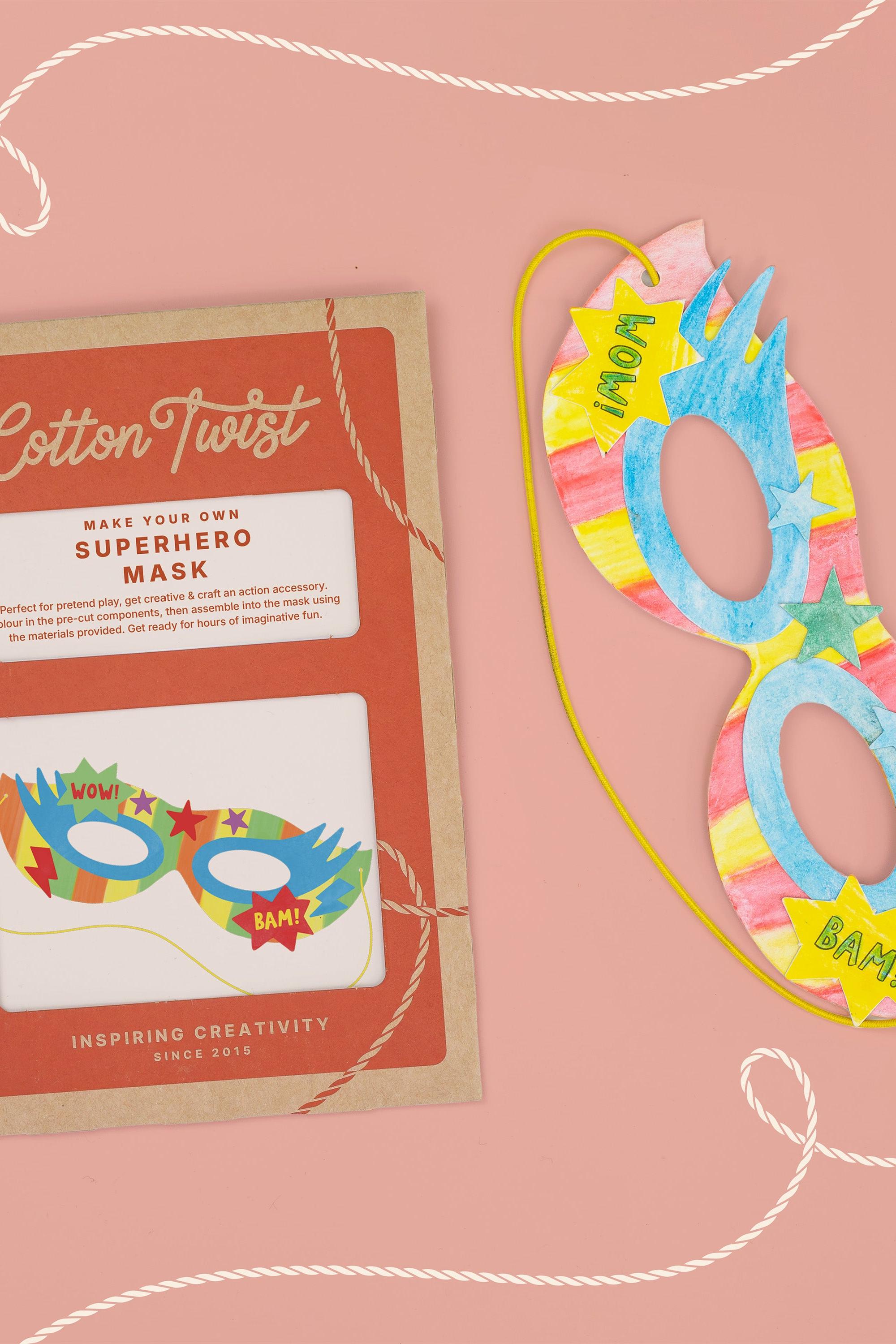 A Cotton Twist DIY superhero mask craft kit next to a completed, hand-colored paper mask.