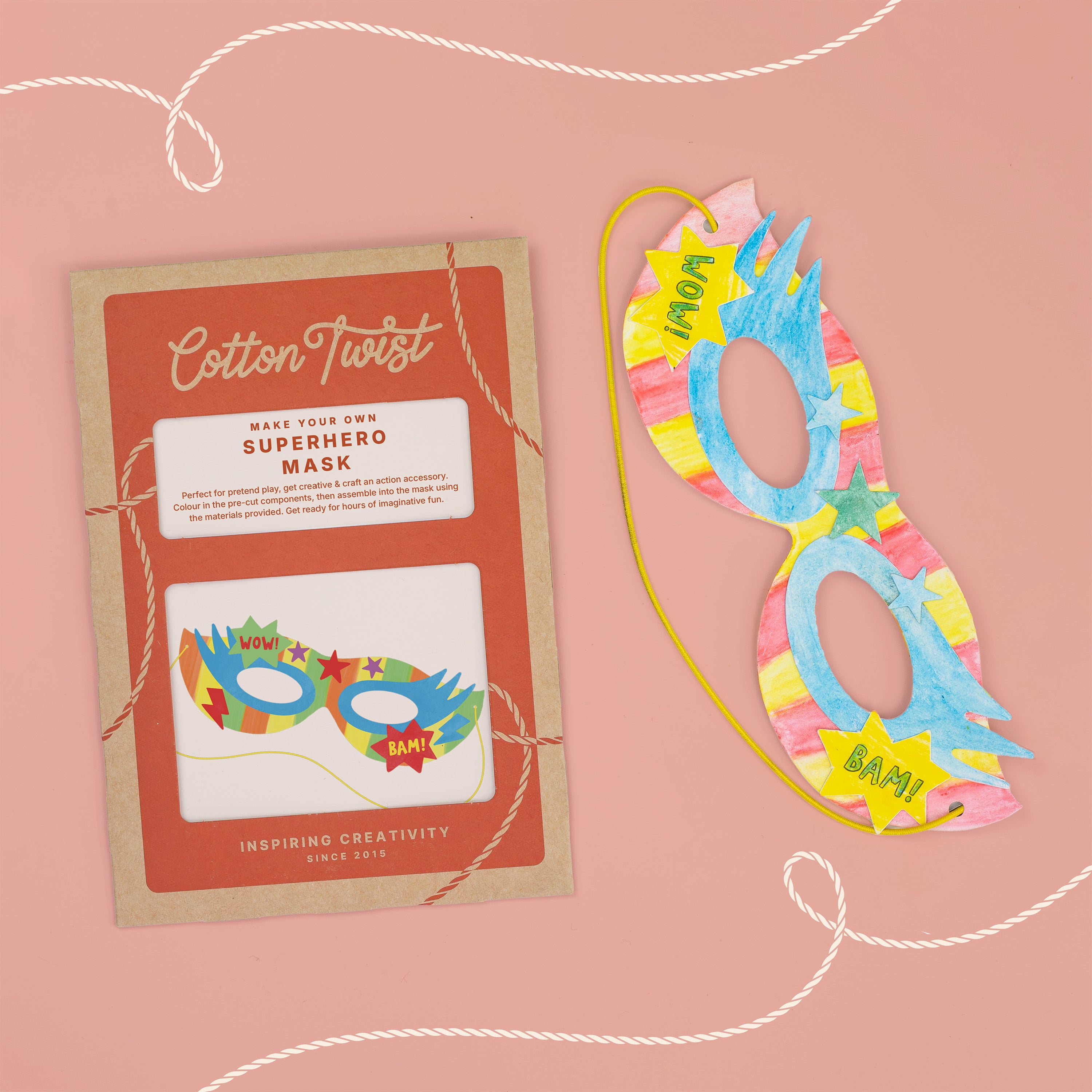 A Cotton Twist DIY superhero mask craft kit next to a completed, hand-colored paper mask.