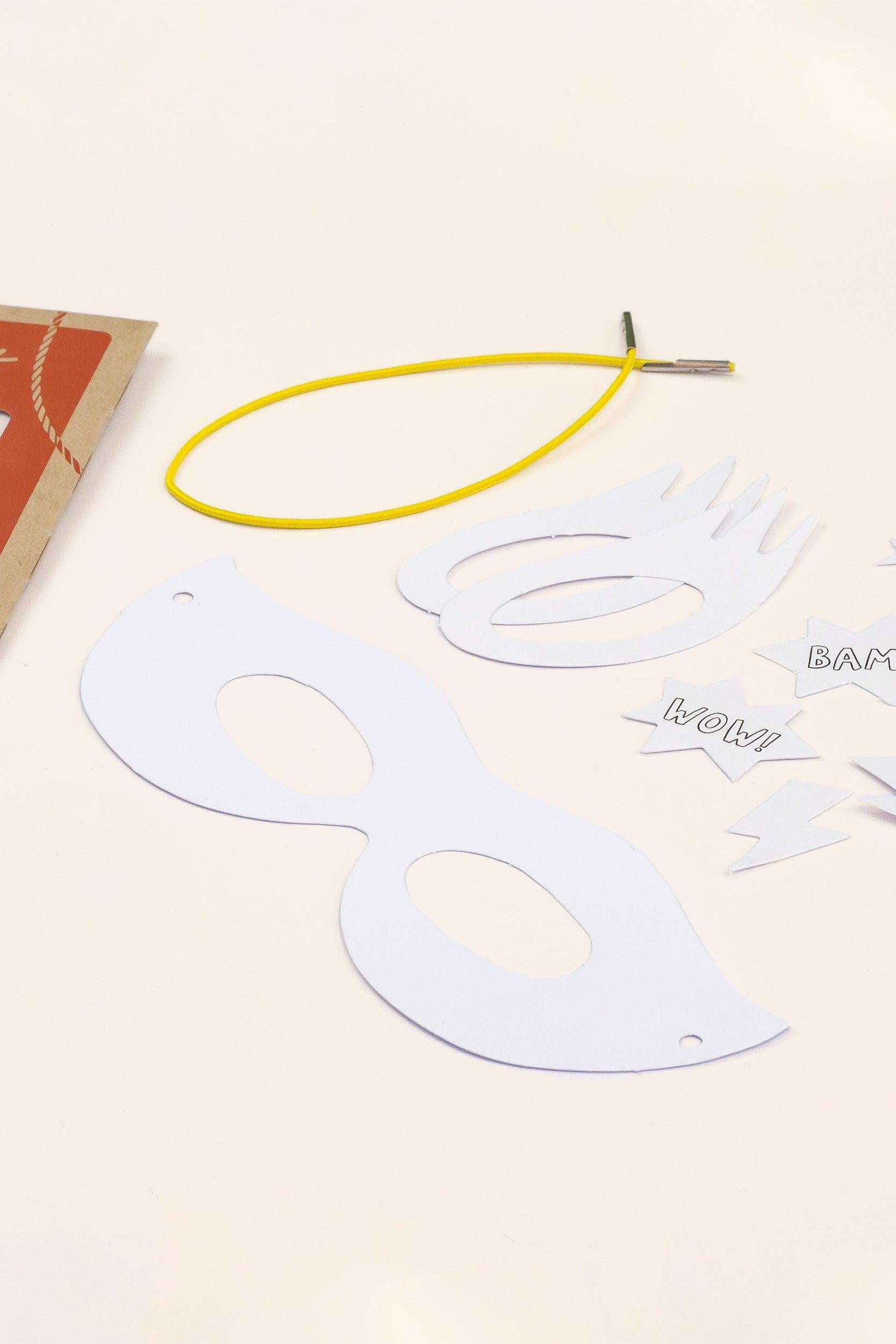 DIY superhero mask craft kit with white paper mask, yellow elastic band, and comic book style cutouts.