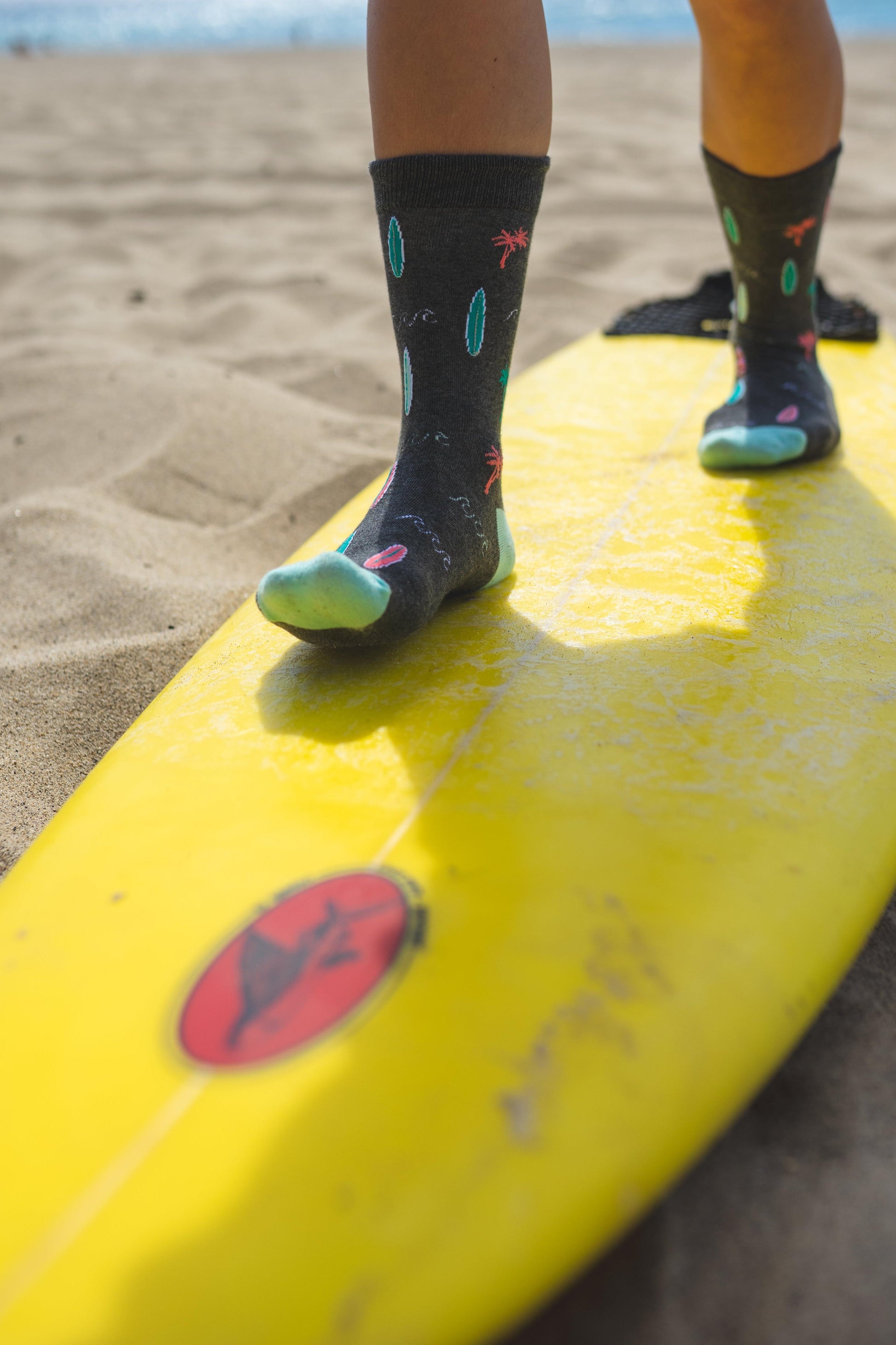 Person standing on a yellow surfboard with feet wearing colorful socks on a sandy beach.