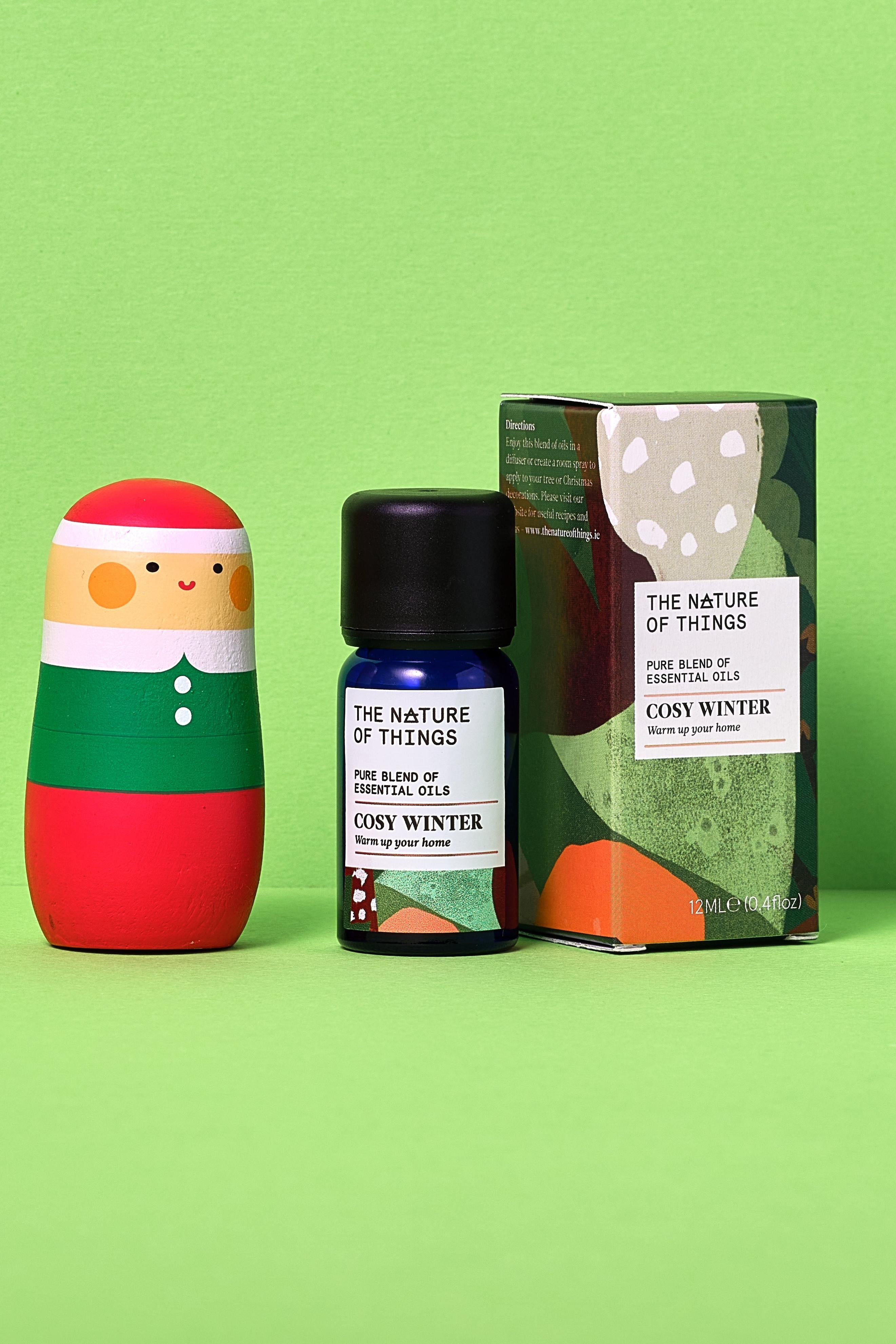 Cosy Winter essential oil bottle, packaging, and colorful elf figurine on a green background