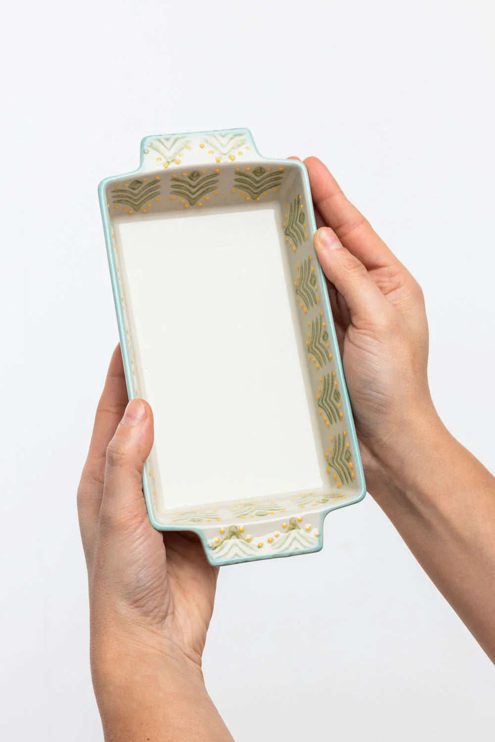 Rectangular ceramic tray with decorative border held by hands on a white background