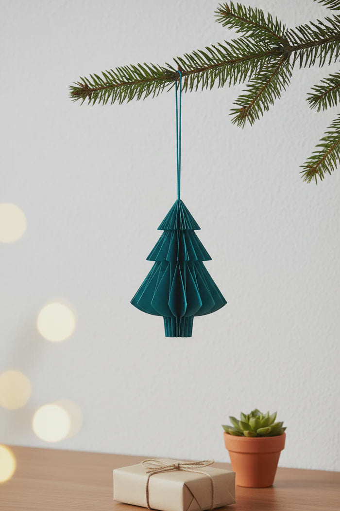 Teal paper Christmas tree ornament hanging from a branch with a gift and plant in the background.