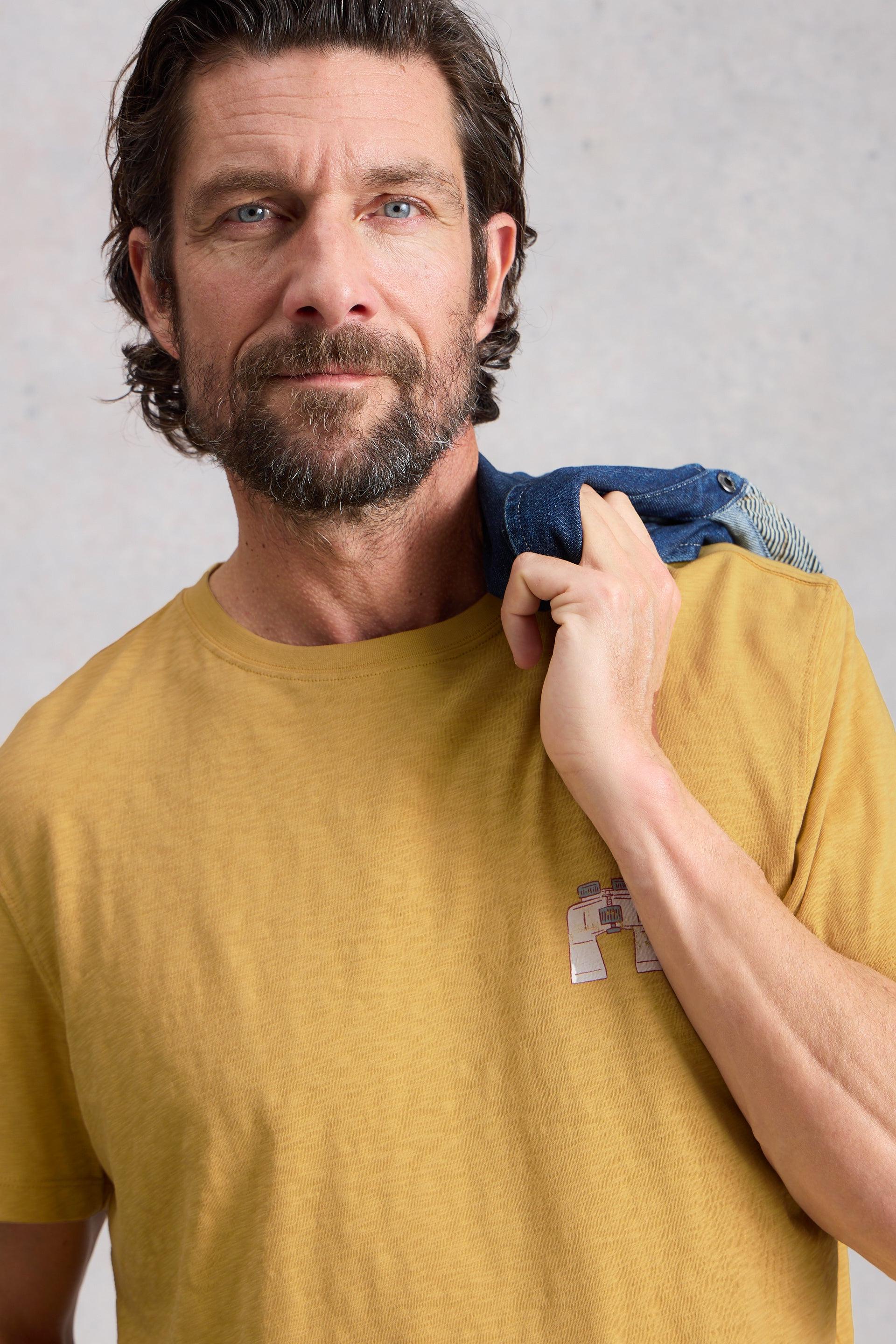 Man wearing a mustard yellow t-shirt with a binoculars design against a plain background