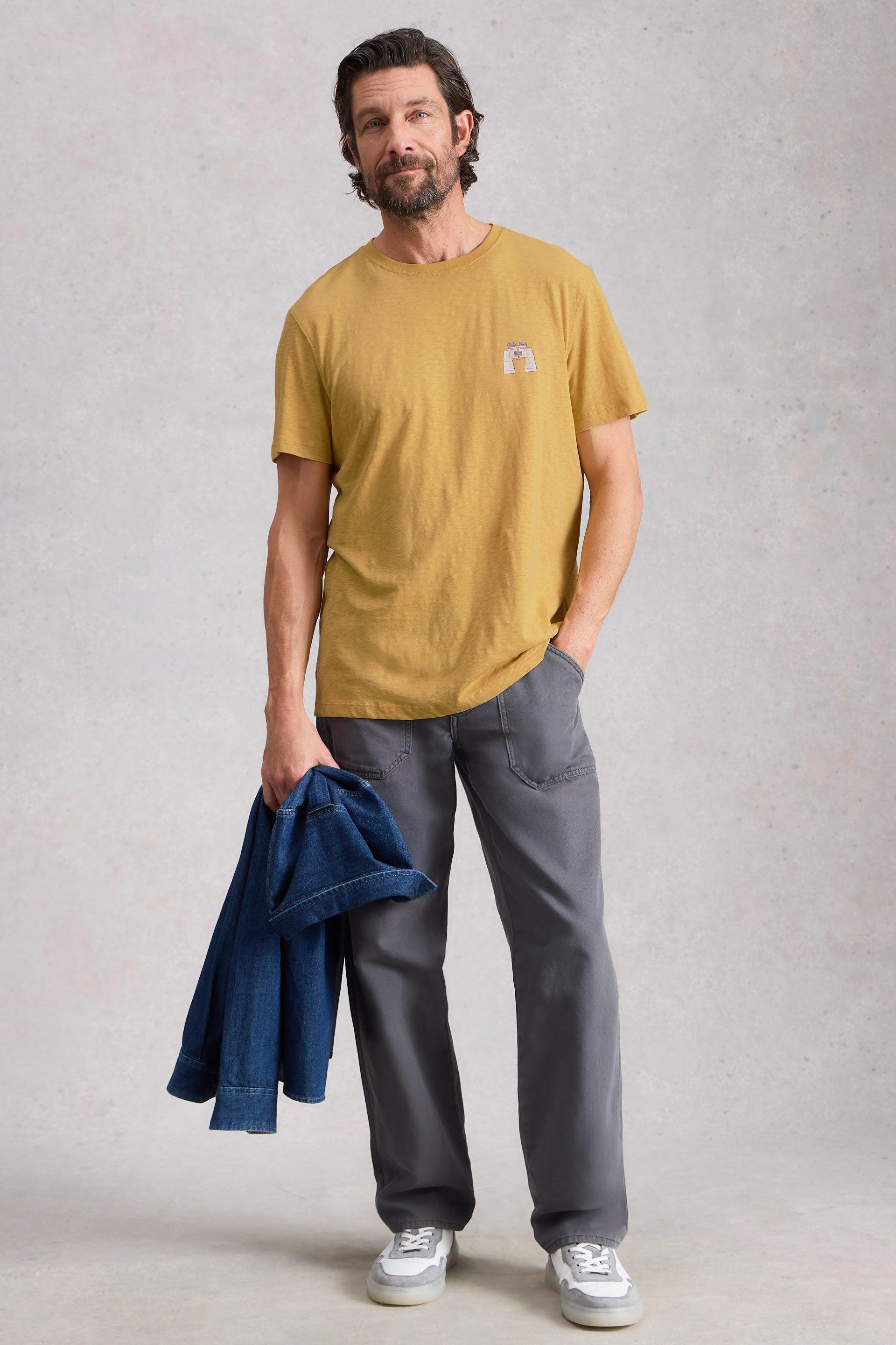 Man wearing a mustard yellow cotton t-shirt and gray pants, holding a blue jacket against a plain background