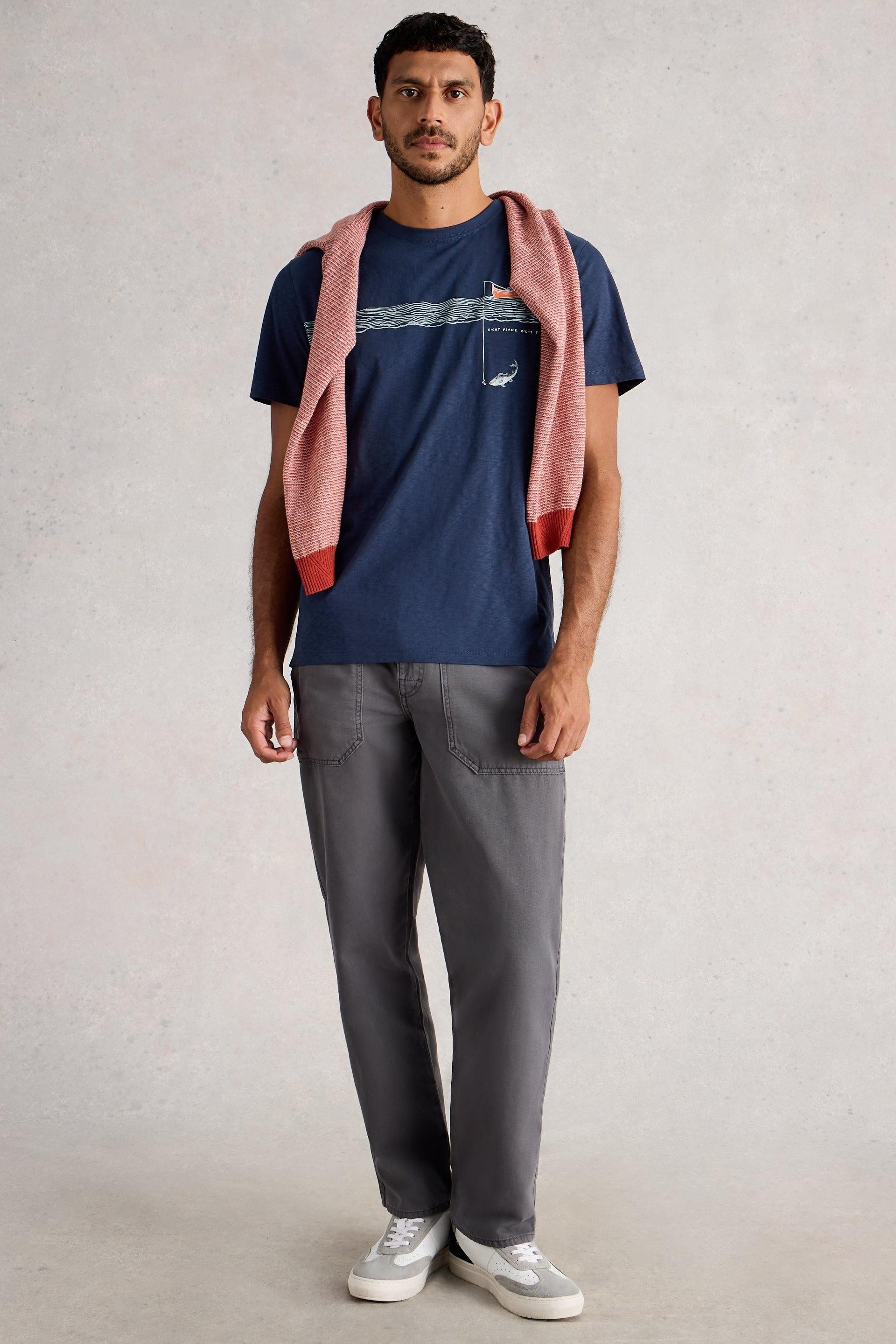 Man wearing a navy t-shirt with a logo, gray pants, and white sneakers, holding a pink sweater.