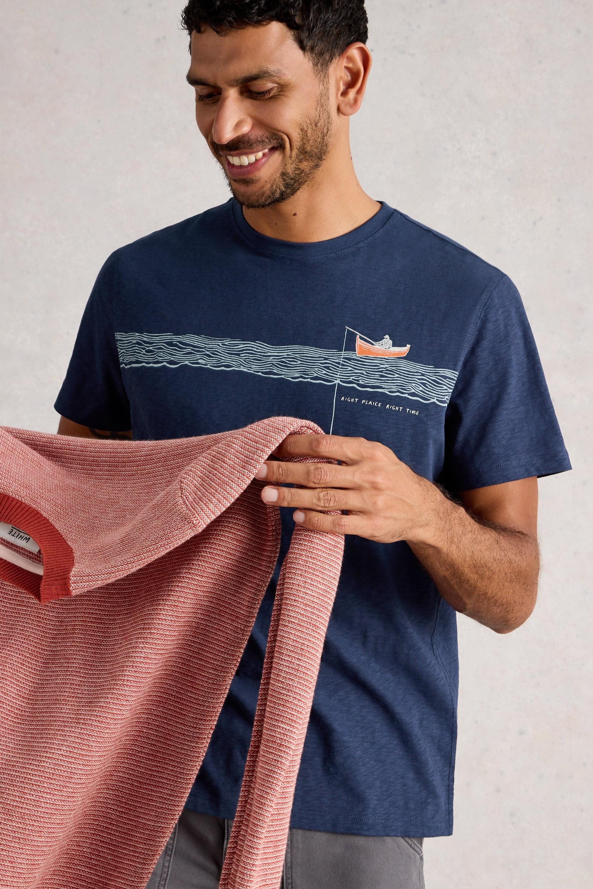Man wearing a navy cotton t-shirt with a graphic design, holding a pink sweater against a light background