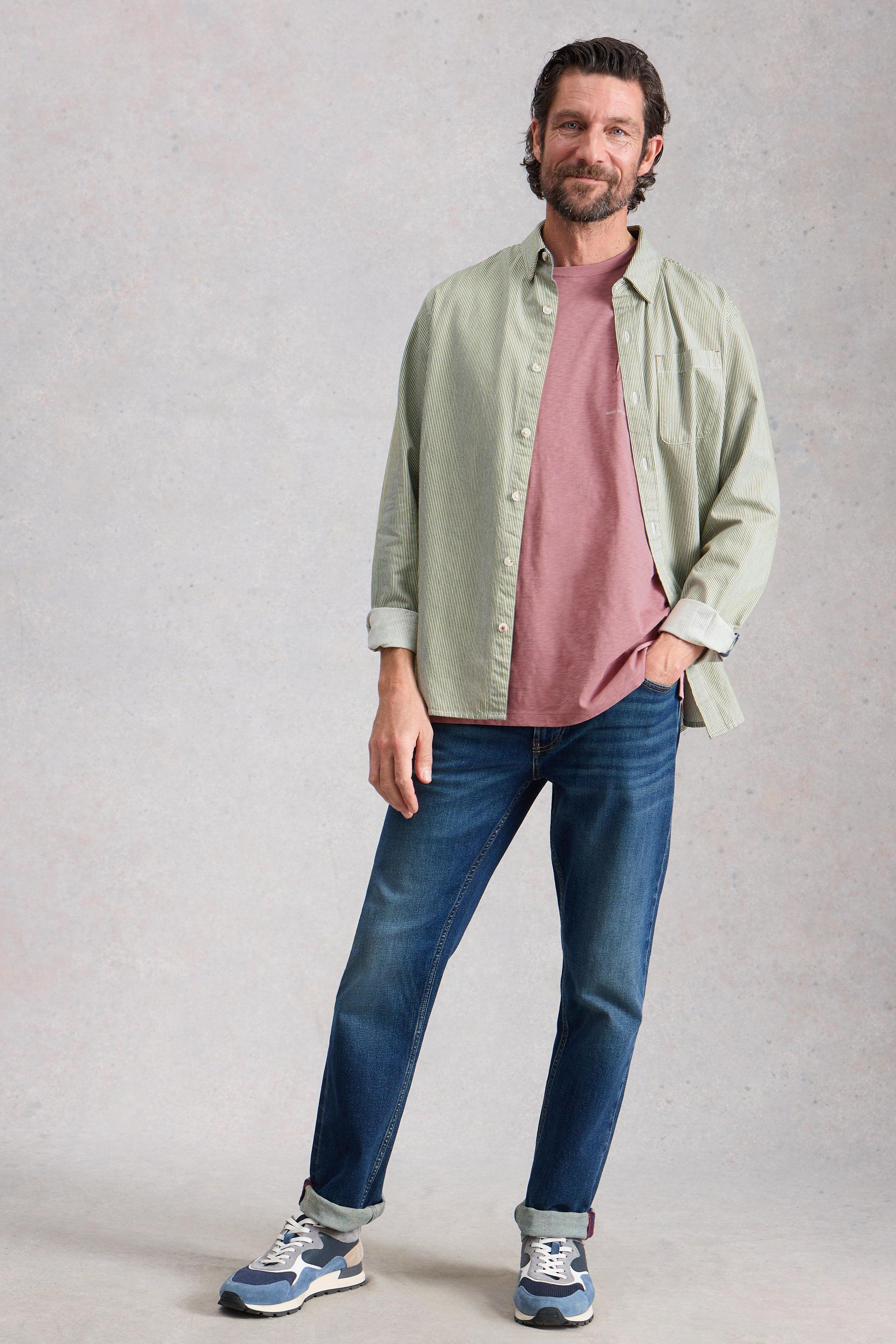 Man wearing a light green shirt over a pink t-shirt, blue jeans, and sneakers on a plain background