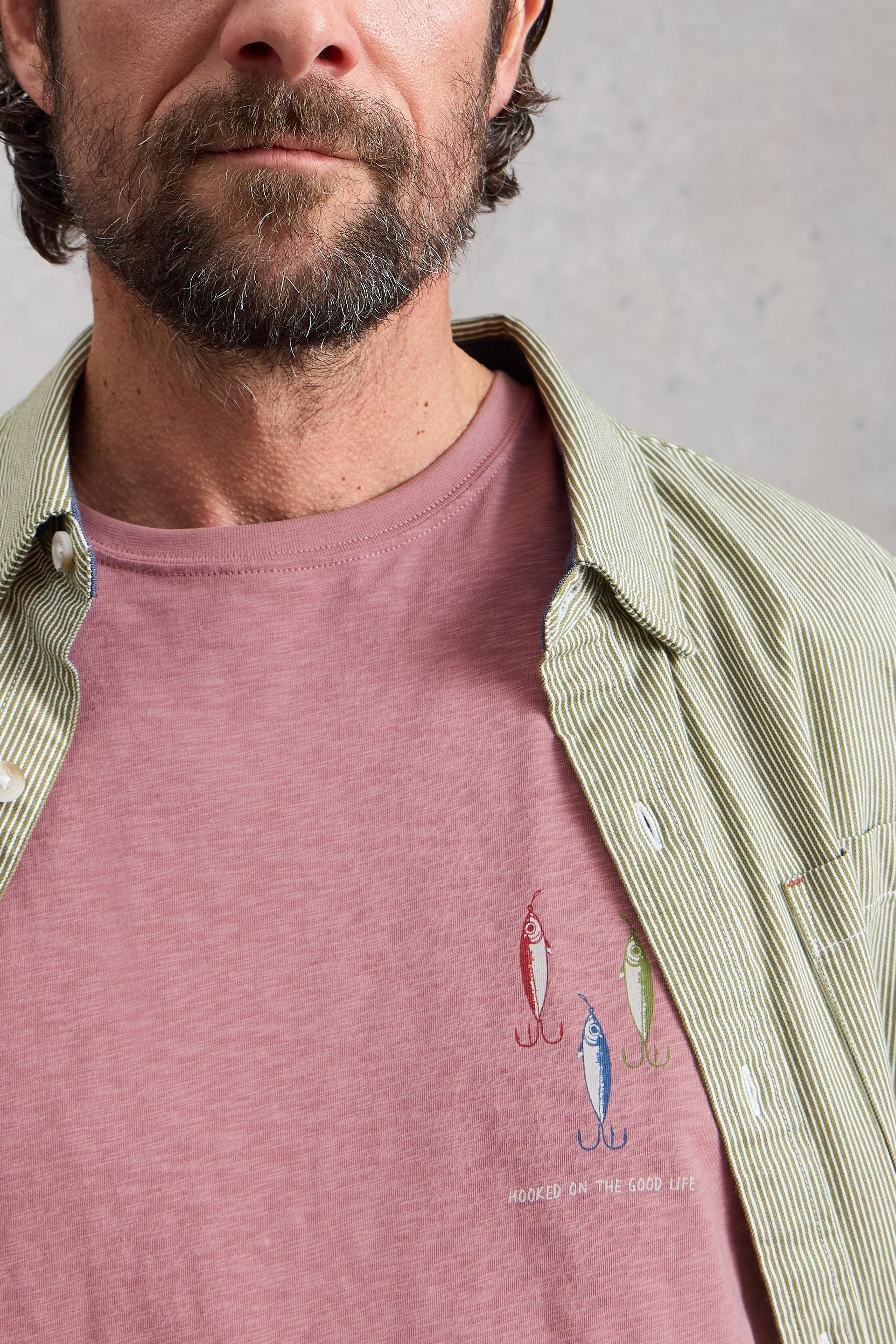 Man wearing a pink cotton t-shirt with fish illustrations and text, paired with a green shirt over a gray background