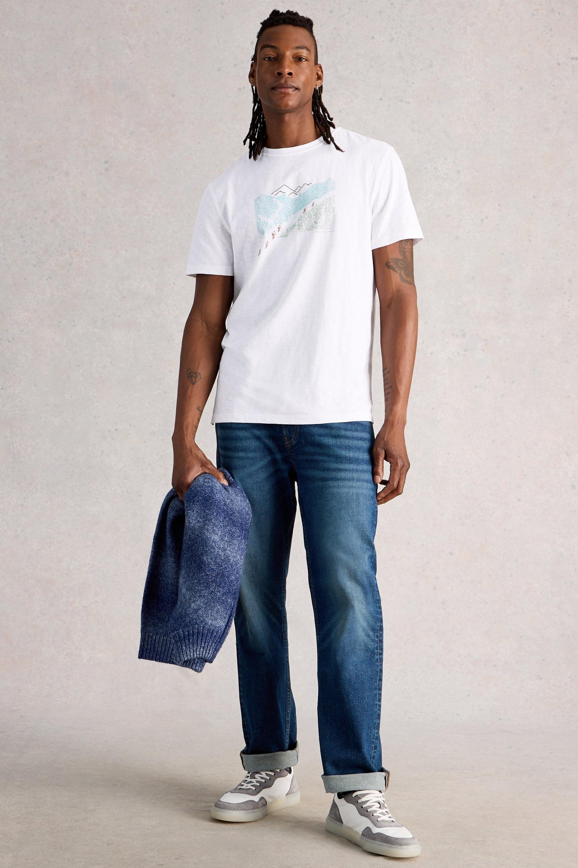 Man wearing a white cotton t-shirt and blue jeans holding a blue sweater against a plain background