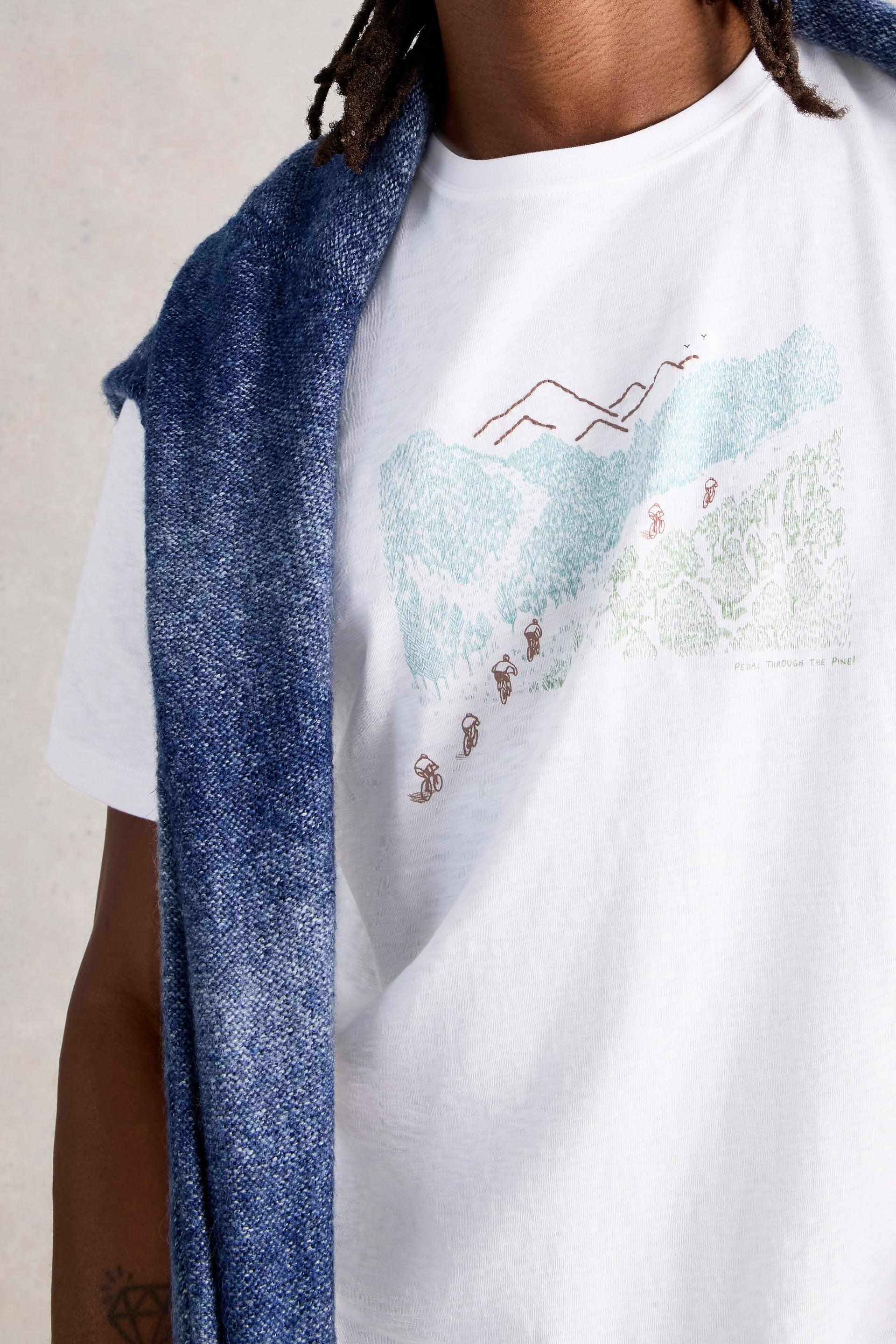 Model wearing a white t-shirt with a graphic design of mountains and people, draped with a blue sweater.