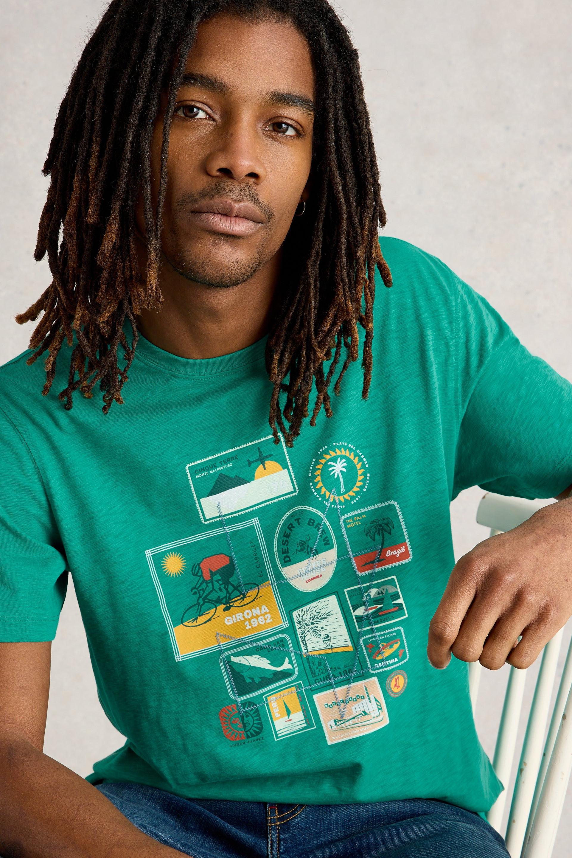 Model sitting on chair wearing teal cotton t-shirt with travel graphic design.