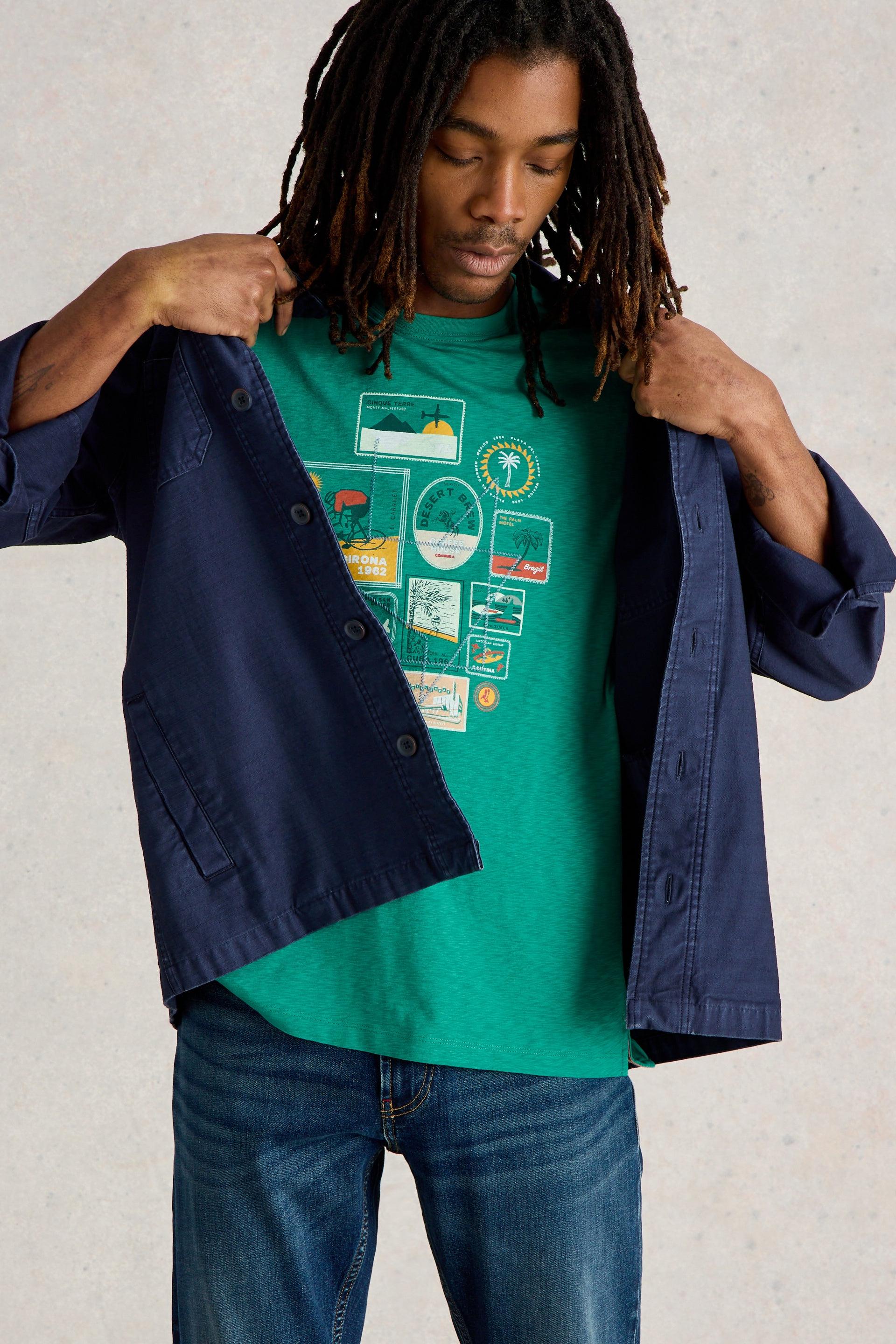 Male model wearing green shirt with travel graphic design layered with a navy shirt.