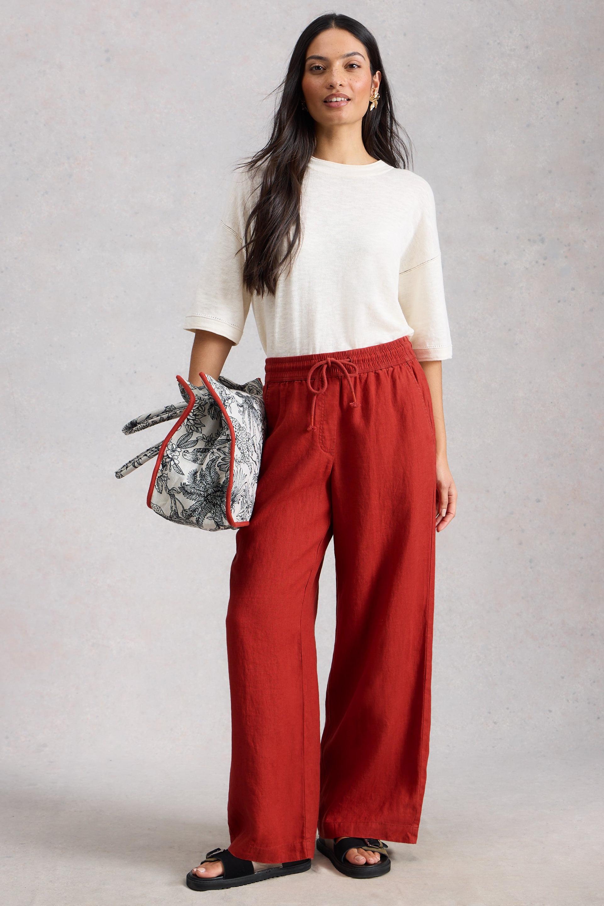 Woman wearing a white top and red wide leg linen pants holding a patterned bag against a gray background